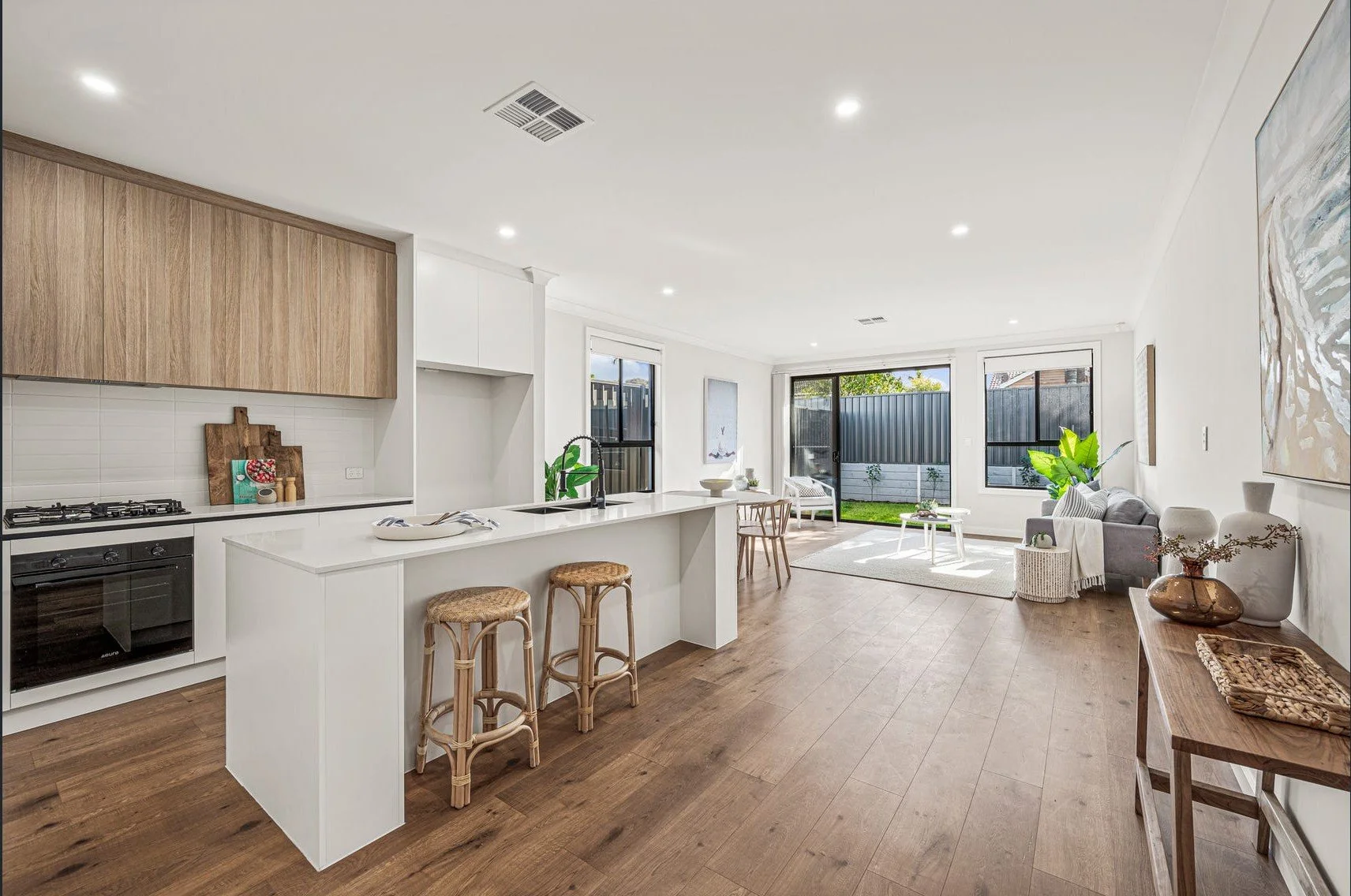 Bright modern open-plan kitchen and living room with island seating, wood cabinets, hardwood floors, and backyard views
