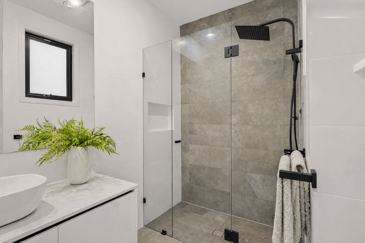 Modern white bathroom with glass-enclosed gray tile walk-in shower, black fixtures, floating vanity, vessel sink, and fern plant