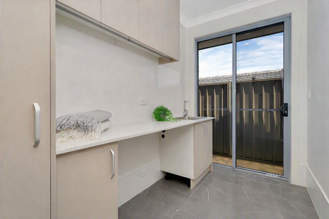 Modern white laundry room with built-in cabinets, countertop, sink, folded blankets, potted plant, and sliding glass door.