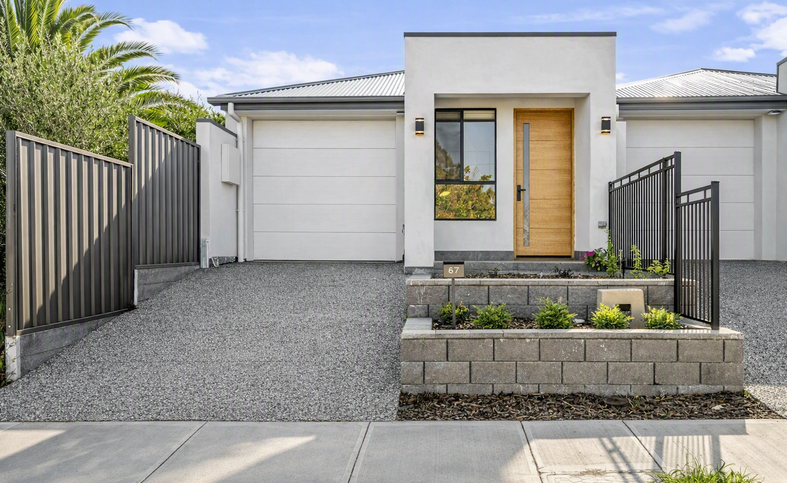 Modern white single-story home with a wooden front door, attached garage, gated driveway, and landscaped raised garden bed.