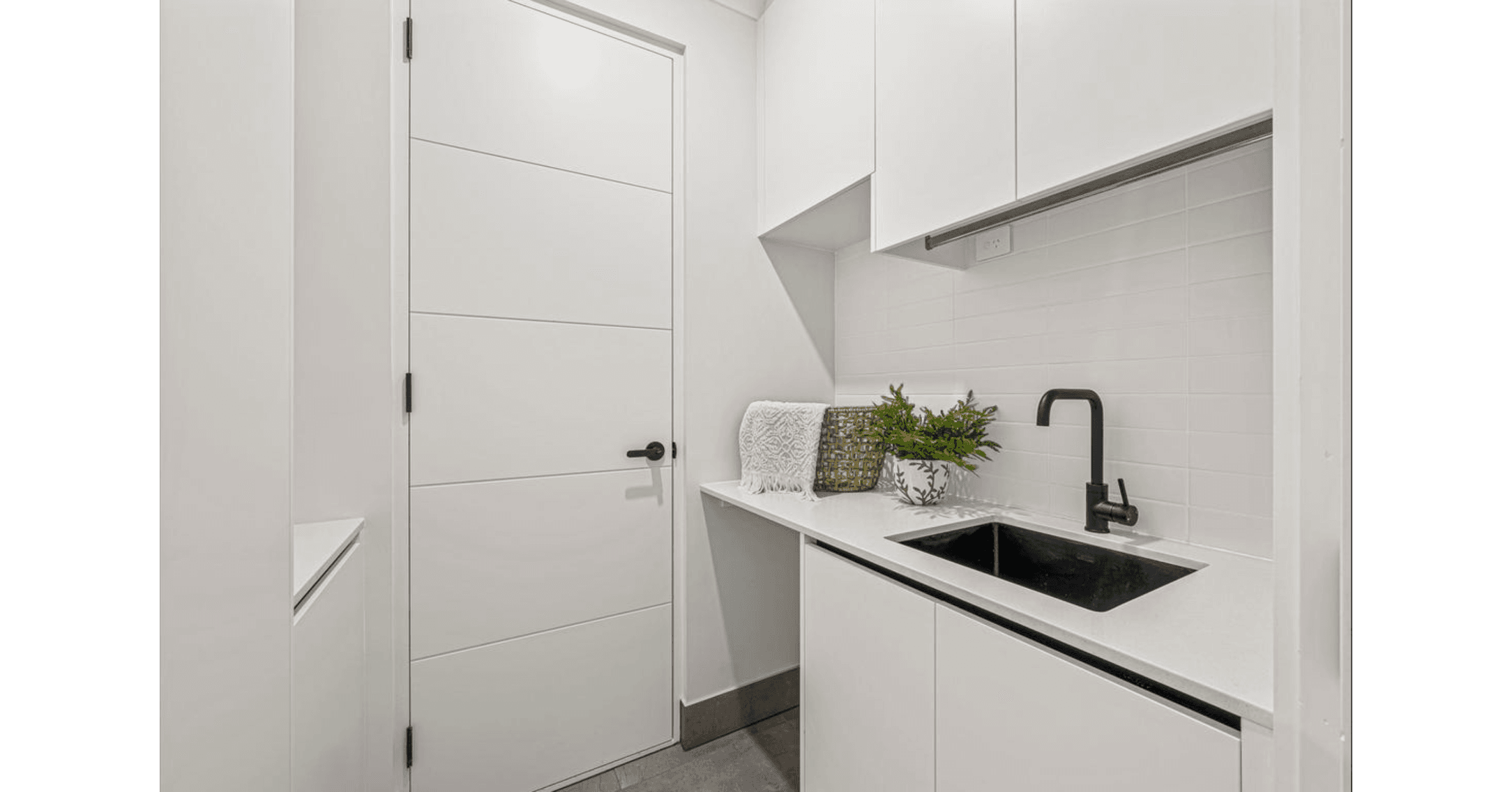 Modern white kitchenette with black sink and faucet, subway tile backsplash, cabinets, and decorative plants