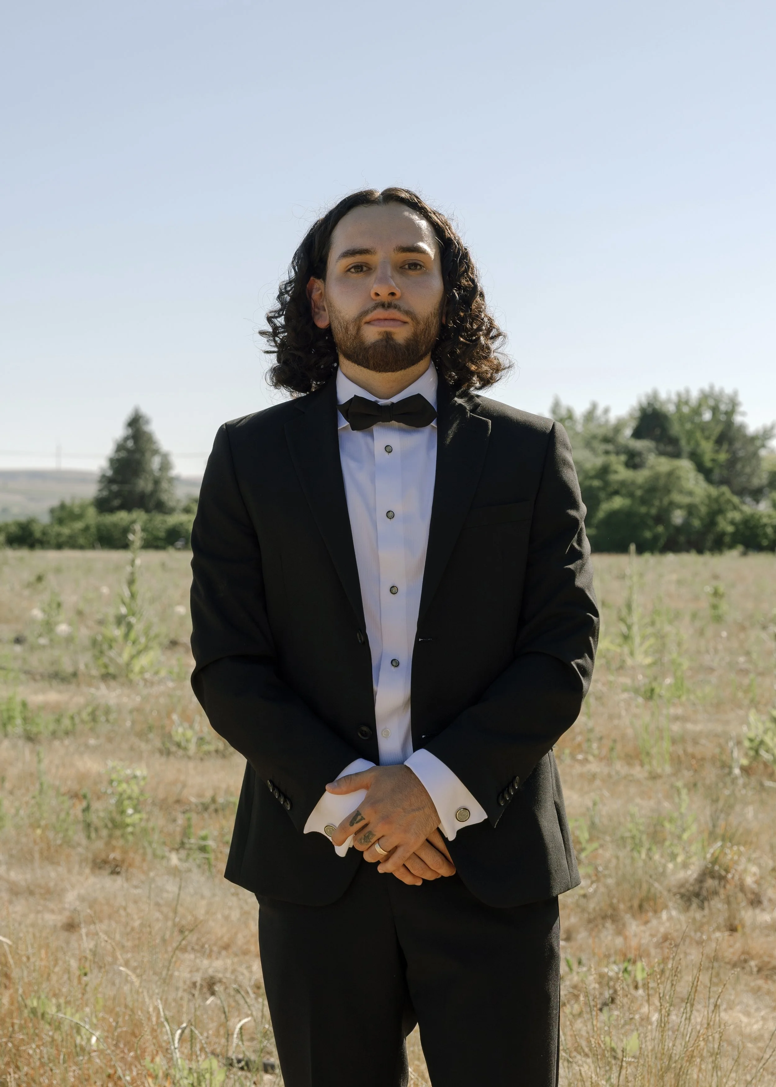 A man with long curly dark hair and a beard is dressed in a black tuxedo with a white shirt and black bow tie, standing outdoors in a field with trees and blue sky in the background.