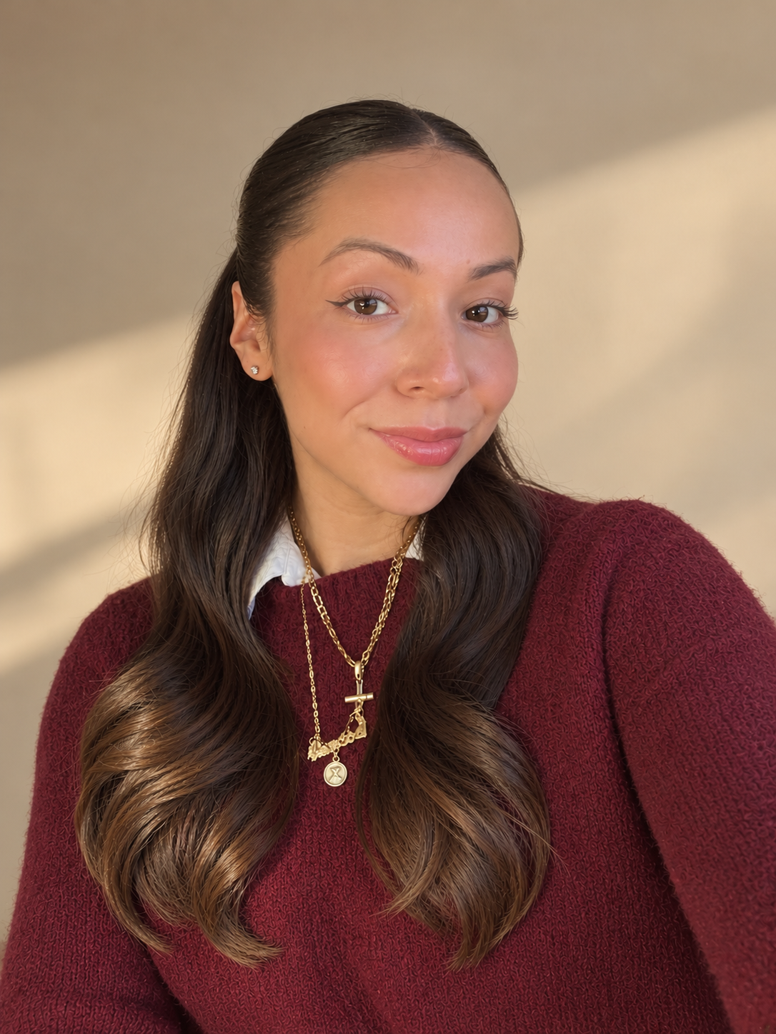 A young woman with long brown wavy hair, wearing a white collared shirt under a red sweater, and layered gold necklaces, taking a selfie against a blurred neutral background.
