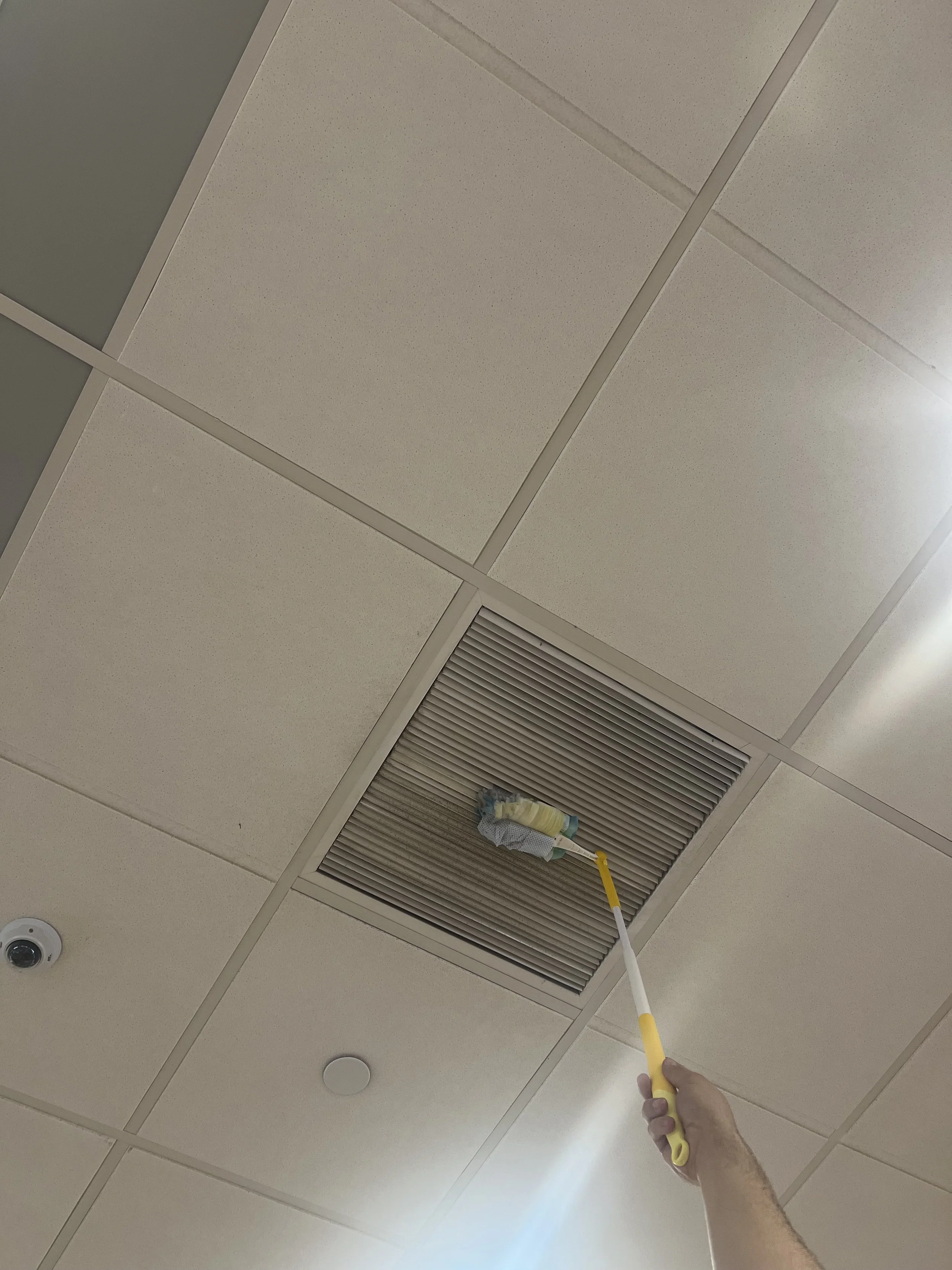 Person cleaning a ceiling vent with a yellow duster using a telescopic tool in an indoor setting.