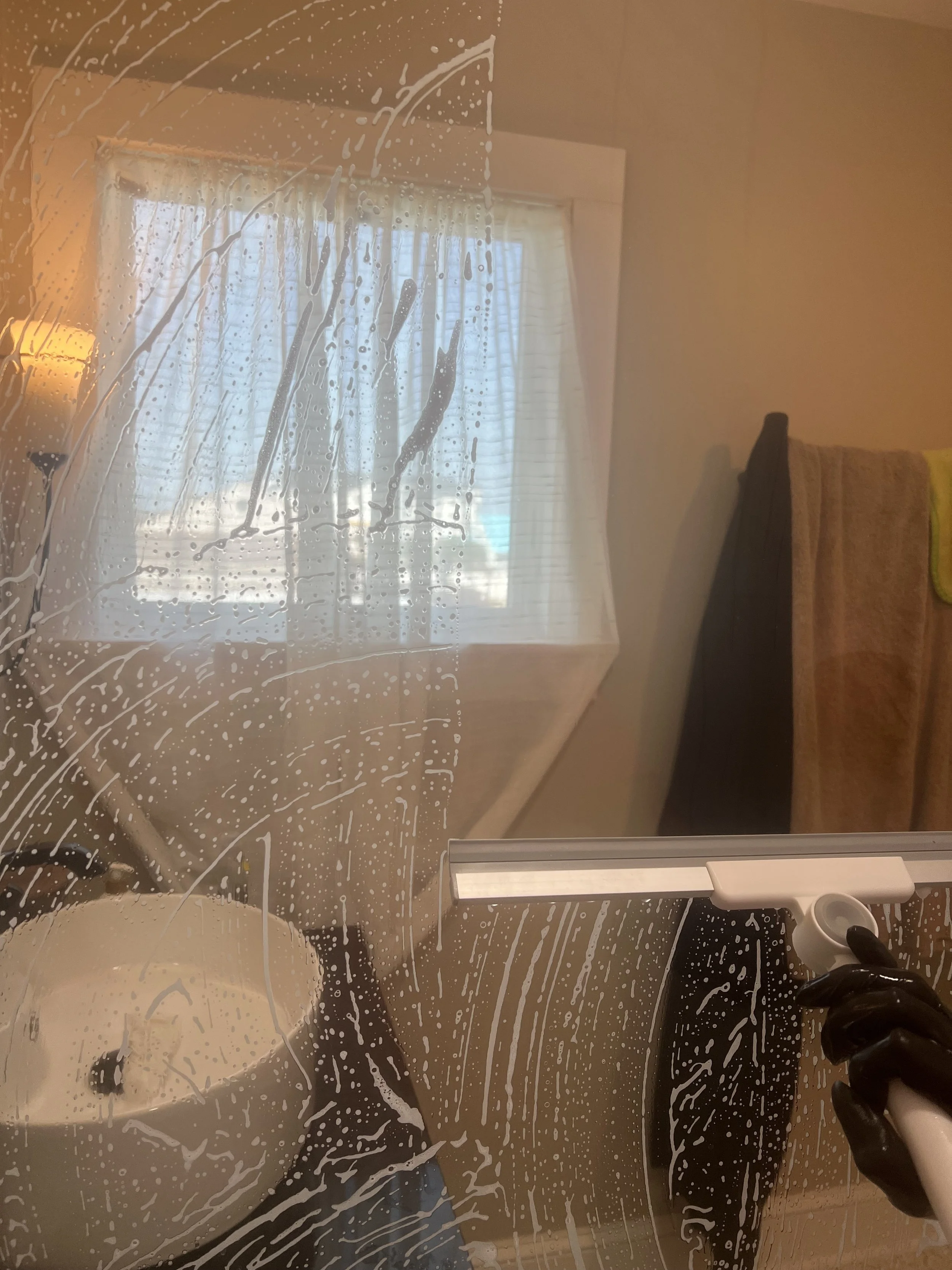 A person cleaning a mirror with a black cloth, with soap or cleaning solution and a vacuum cleaner visible. The mirror has soap suds on it, and the room reflected in the mirror includes a window with blinds, a lamp, and part of a bed with a headboard.