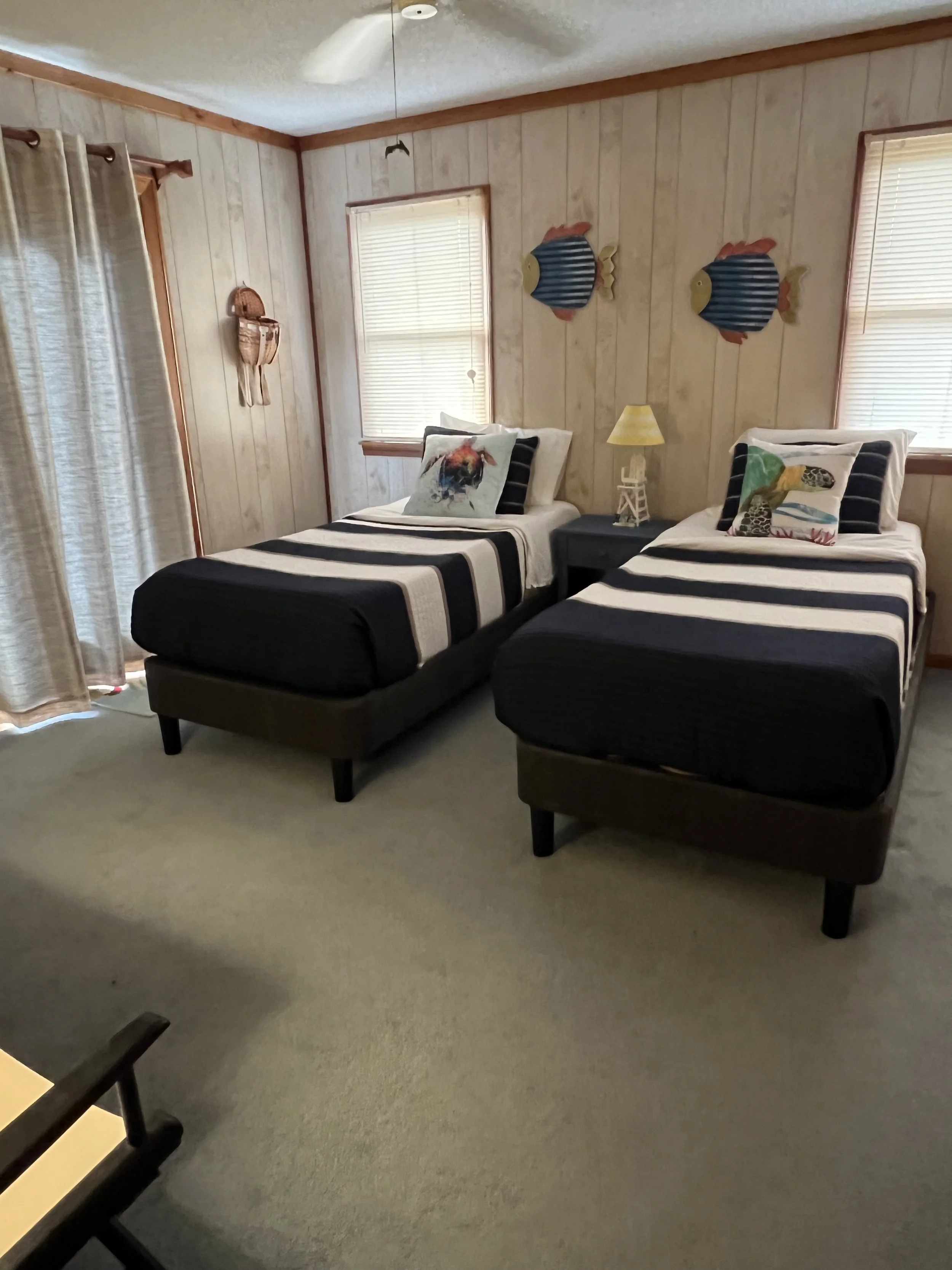 A bedroom with two twin beds, each decorated with safari-themed pillows. The wall features fish wall decorations, and there is a nightstand with a lamp between the beds. Light comes through the window with blinds, and the room has wood-paneled walls.