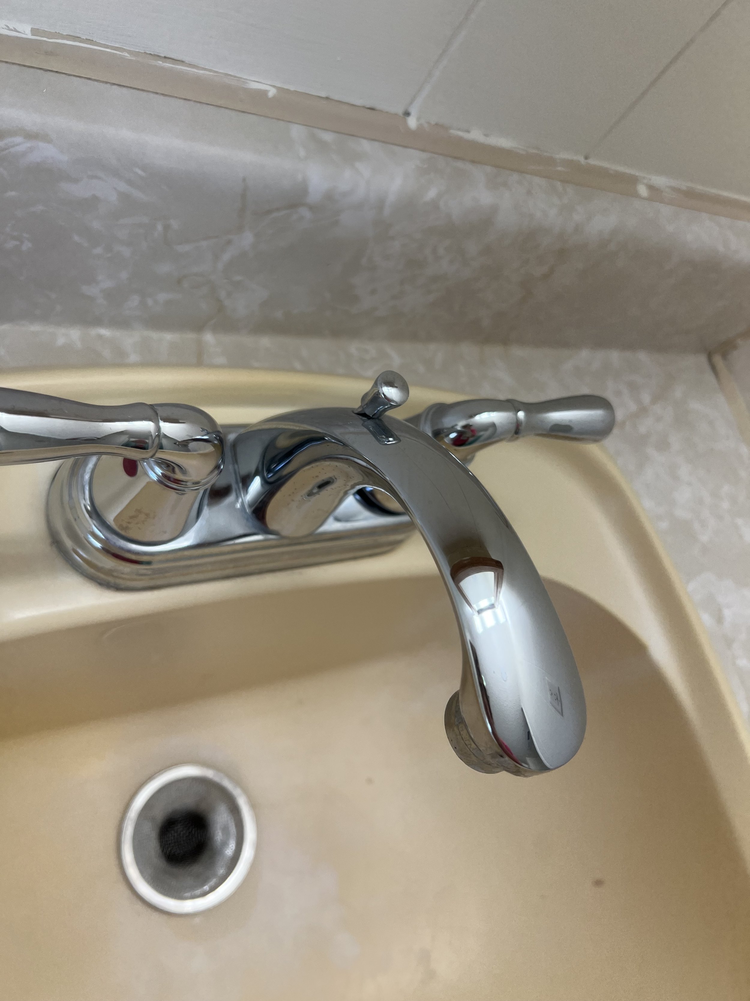 Close-up of a silver faucet with a single curved handle in a bathroom sink with a beige countertop and a drain.