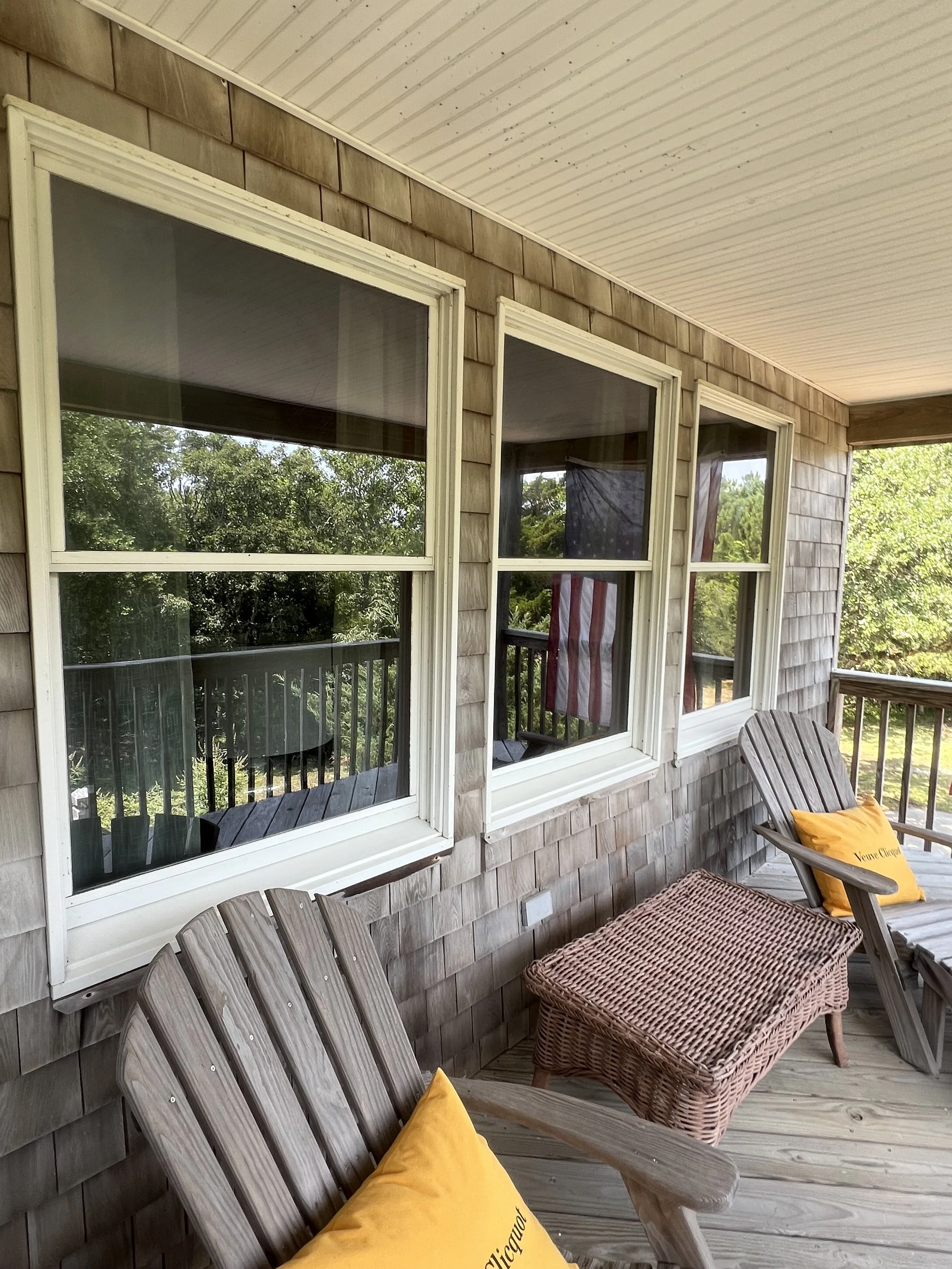 View of a porch with wooden Adirondack chairs and wicker furniture, featuring yellow and orange pillows, overlooking trees and greenery.