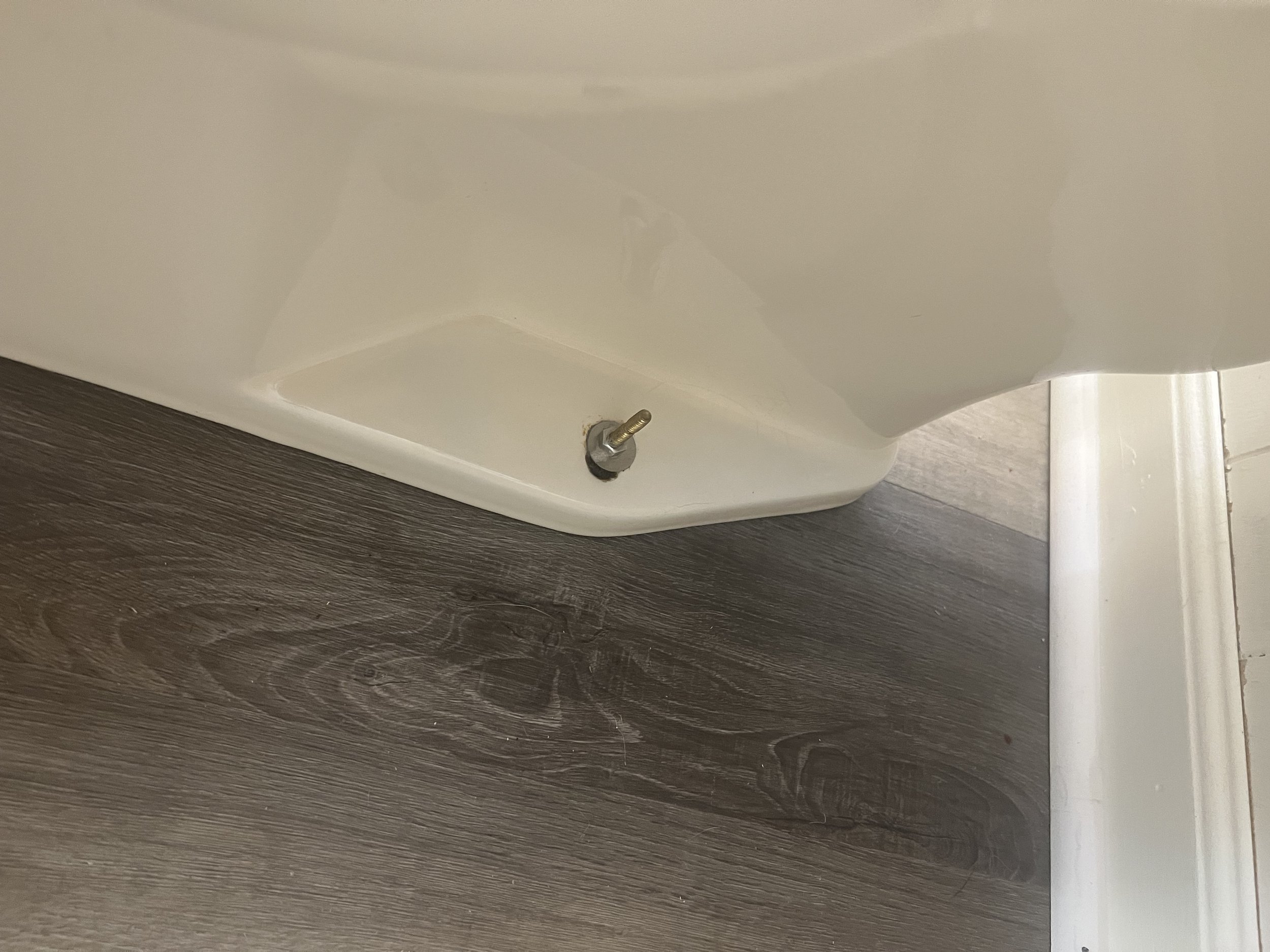 The image shows the base of a white piece of furniture, possibly a table or a desk, with a visible metal bolt installed in the wood floor.