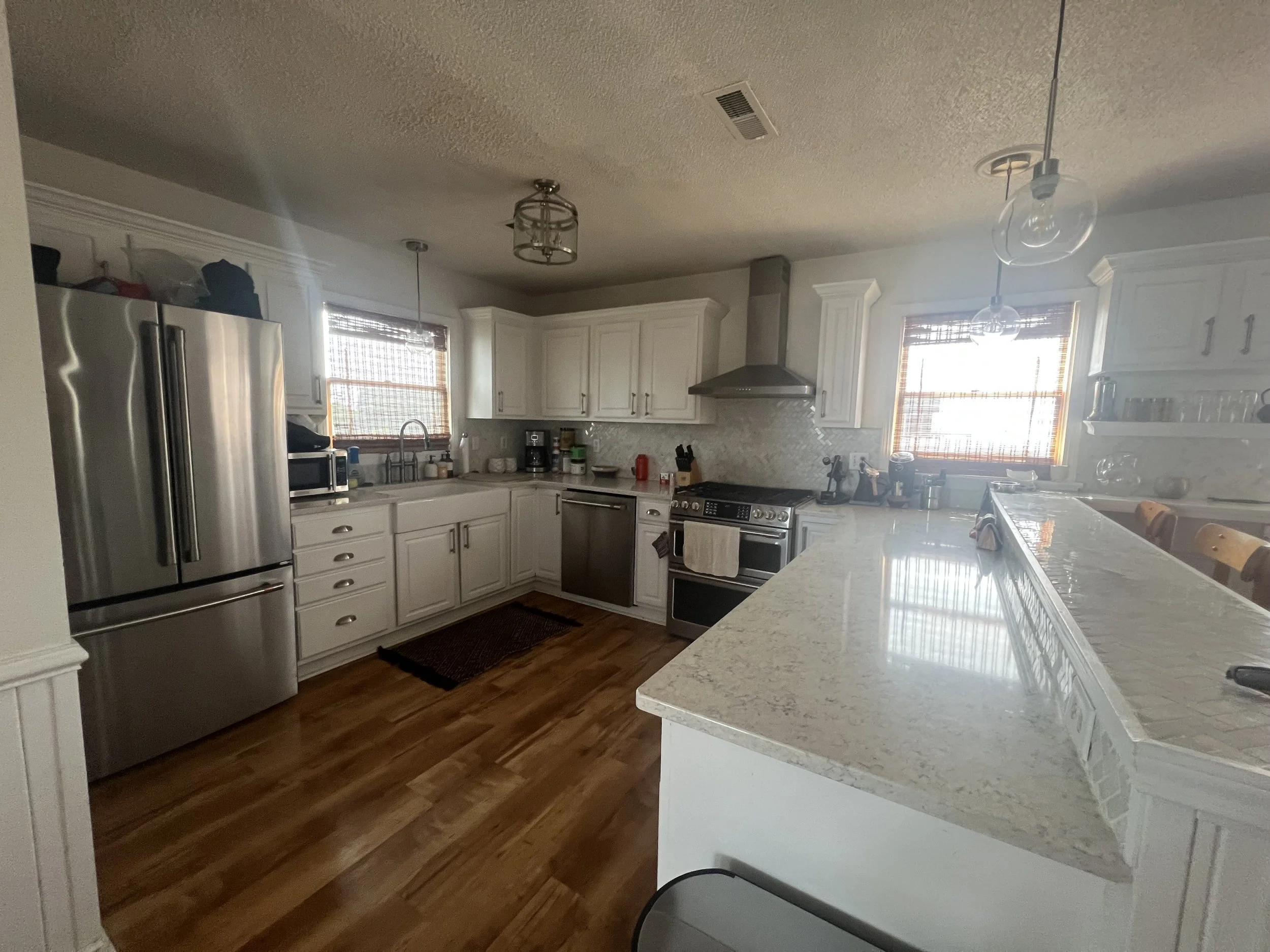 A spacious kitchen with white cabinets, a stainless steel fridge, a stove, and a granite countertop island. Two windows with wooden blinds let in natural light, and various kitchen items are arranged on the counters.