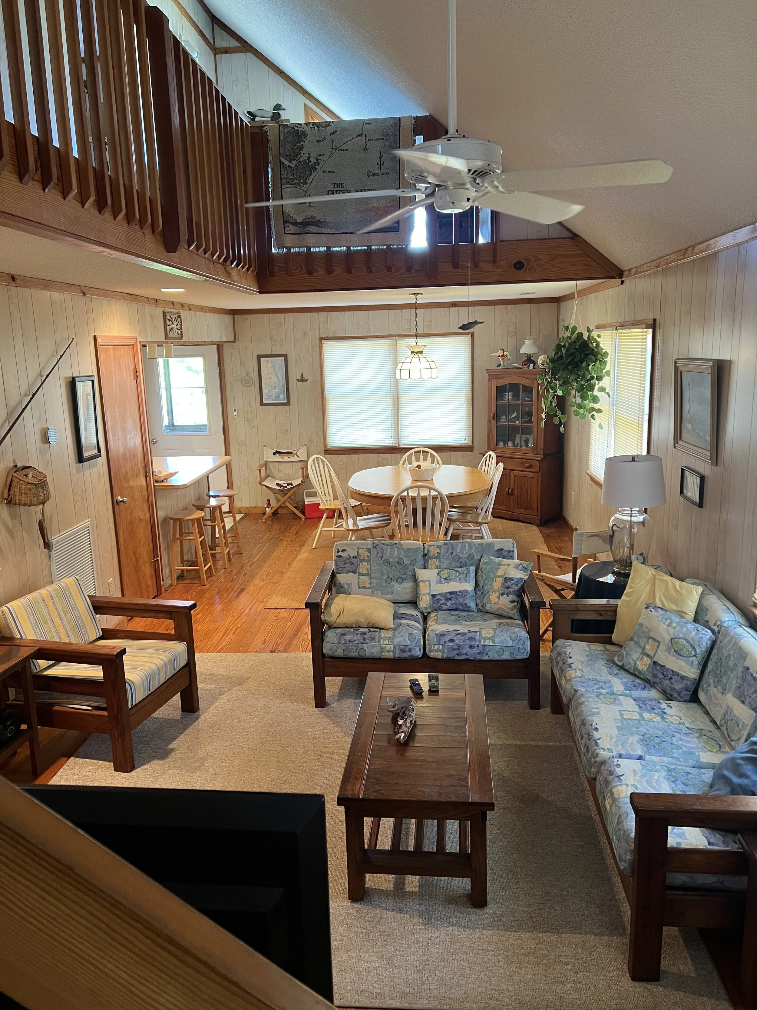 Living room with wooden walls and furniture, a ceiling fan, a dining table with chairs, a sofa, a TV tray table, and large windows with blinds.