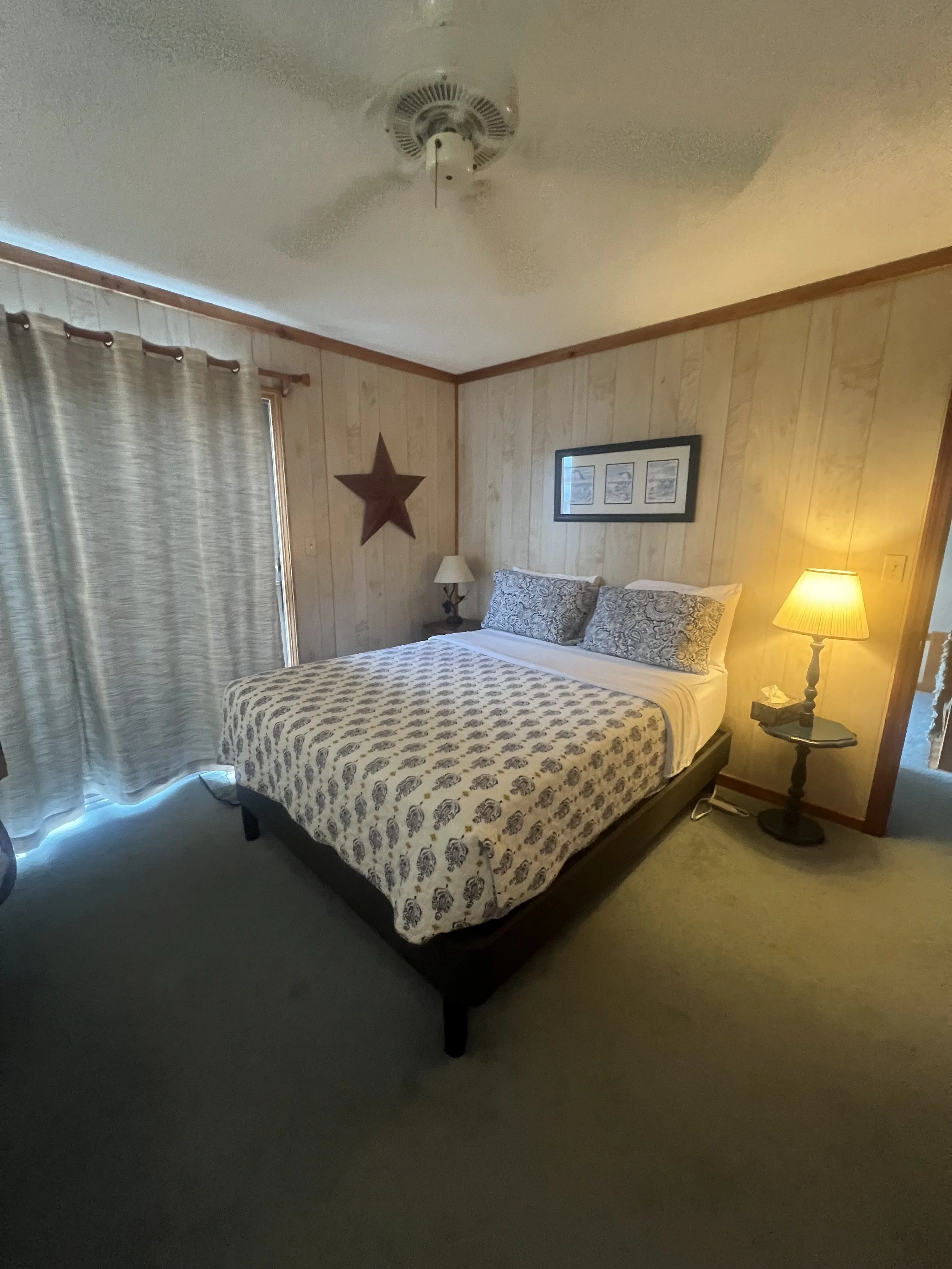 Bedroom with a bed covered in a patterned comforter, two pillows, a nightstand with a lamp, a framed picture on the wall, a wooden star decor, a window with curtains, and a ceiling fan.