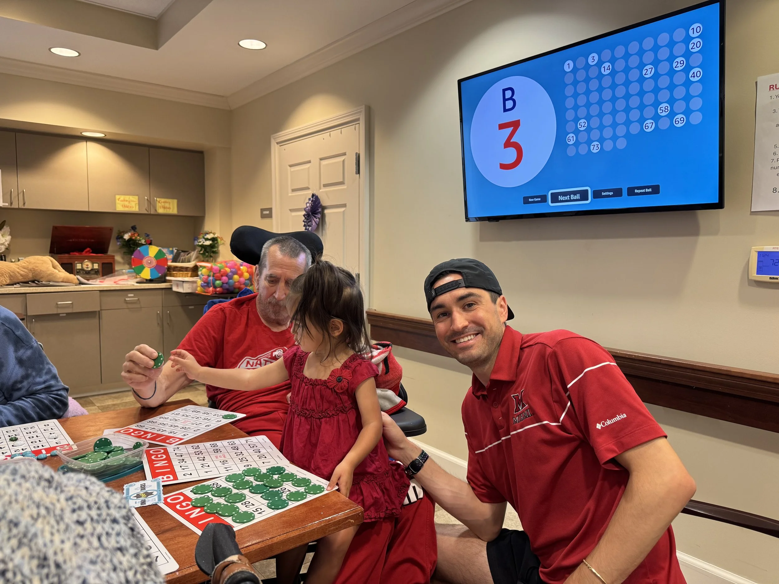 Nathan, his daughter, and his father playing bingo at a table with bingo cards and chips, while a bingo game is displayed on a wall-mounted screen.
