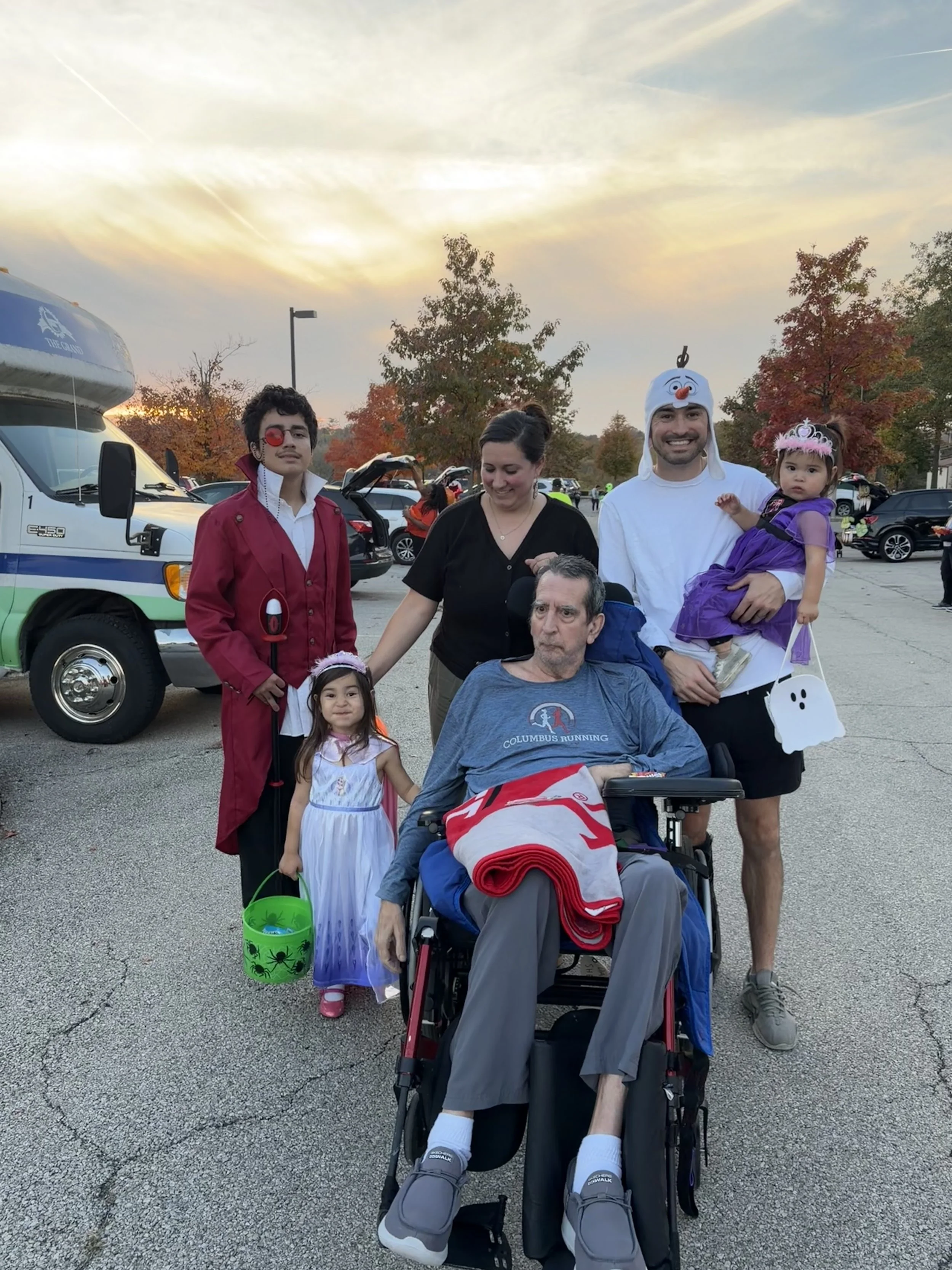 Nathan and his family in Halloween costumes posing outdoors beside a bus at sunset.