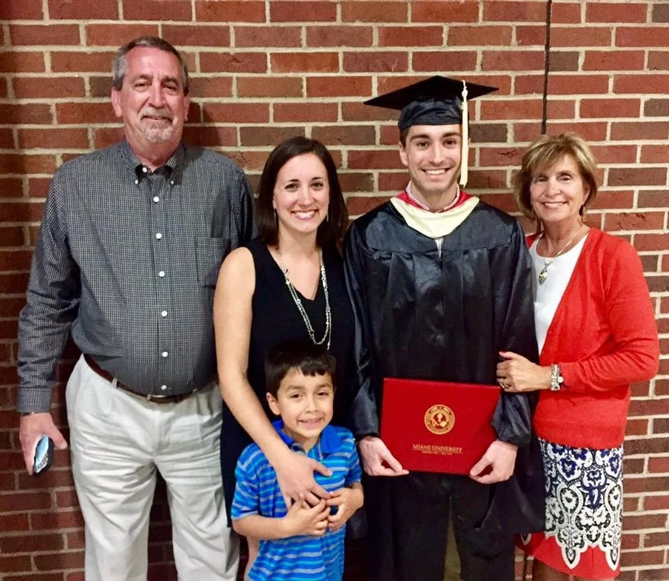 Nathan and family posing in front of a brick wall with a graduate wearing a cap and gown and holding a Miami University diploma folder.