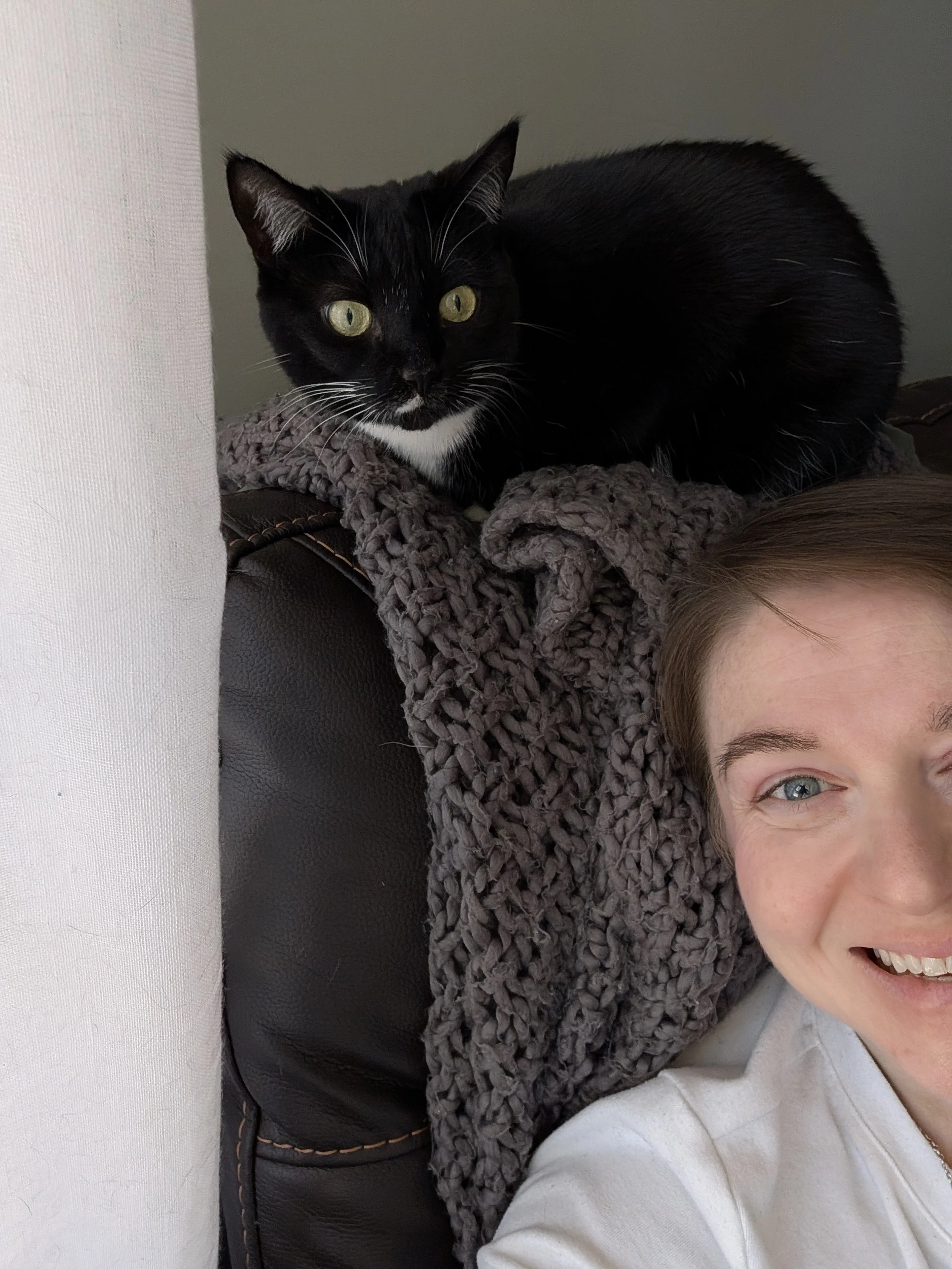 A person with brown hair and blue eyes taking a selfie while a black and white cat is lying on a gray blanket next to them. The person's face is partly visible, showing a big smile.