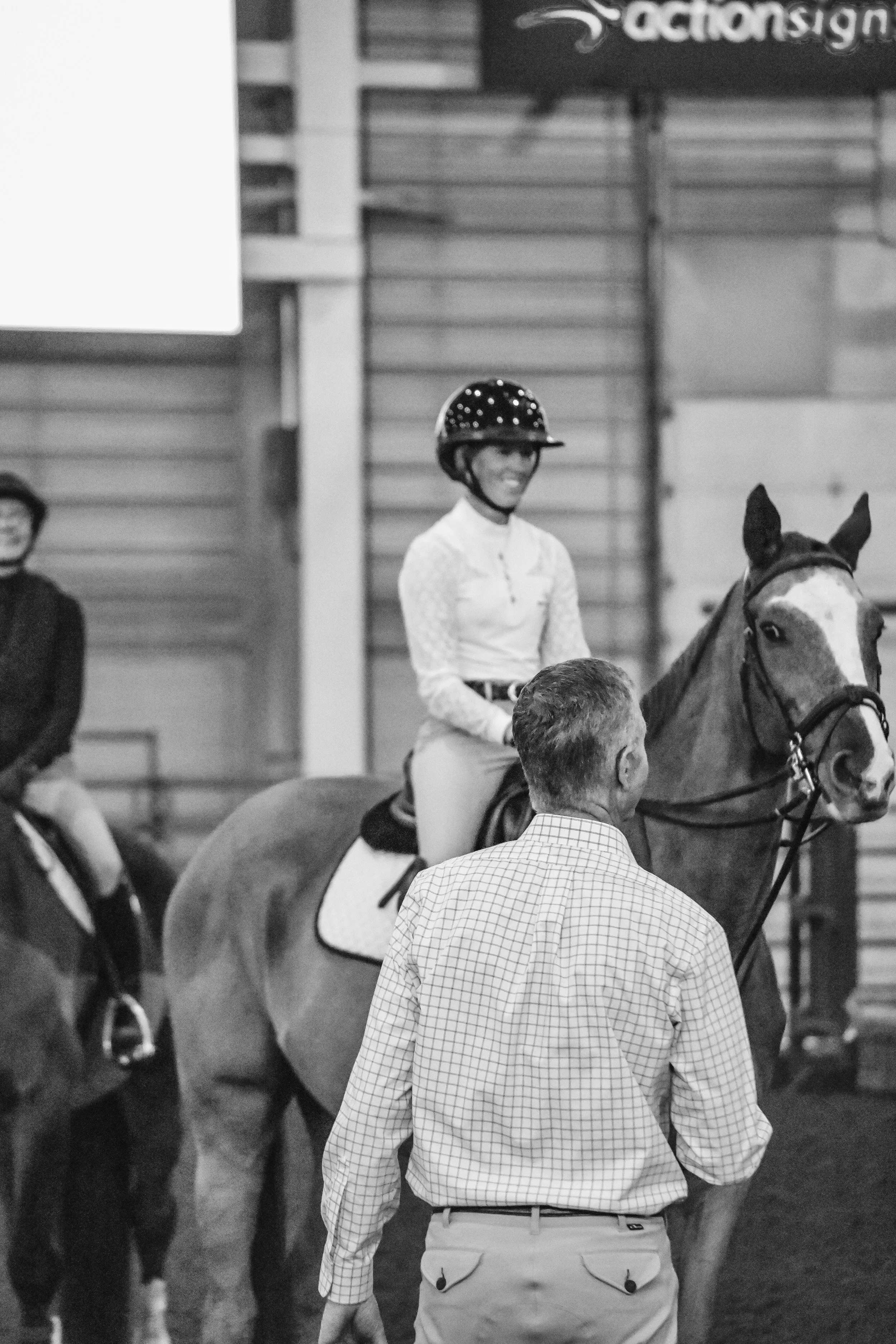 A person wearing a helmet and riding a horse is smiling at an indoor equestrian event. There are spectators and event signs visible in the background.
