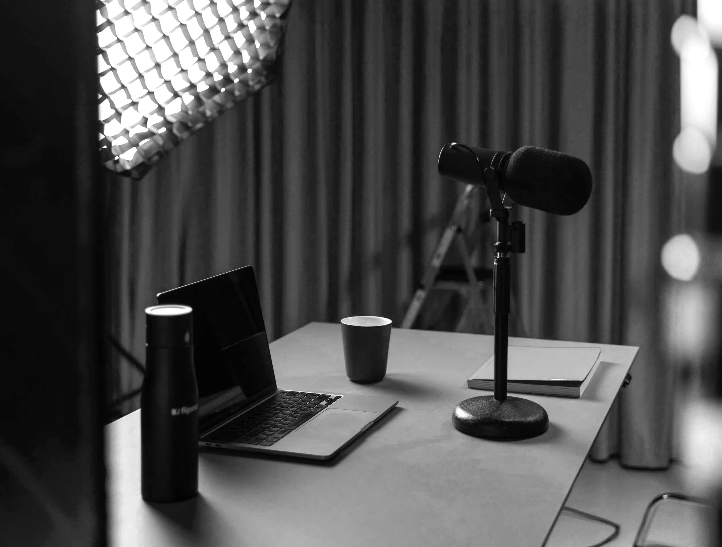 Black-and-white podcast recording setup with a microphone, laptop, coffee cups, books, and studio lighting on a desk.