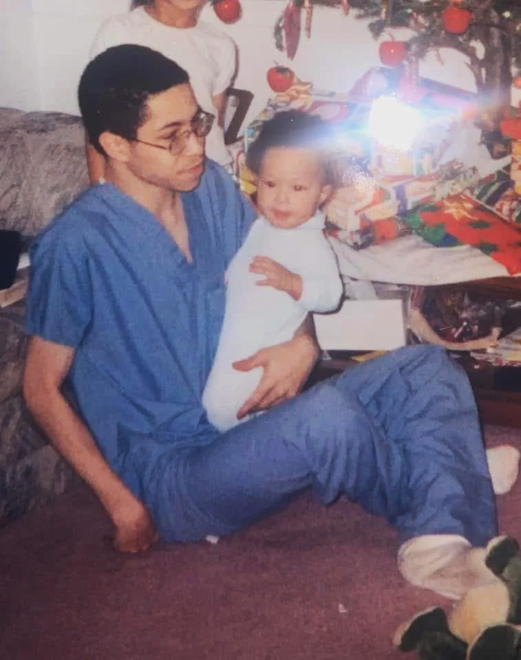Bree Stephens father in blue medical scrubs holding Bree Stephens beside a decorated Christmas tree and wrapped gifts