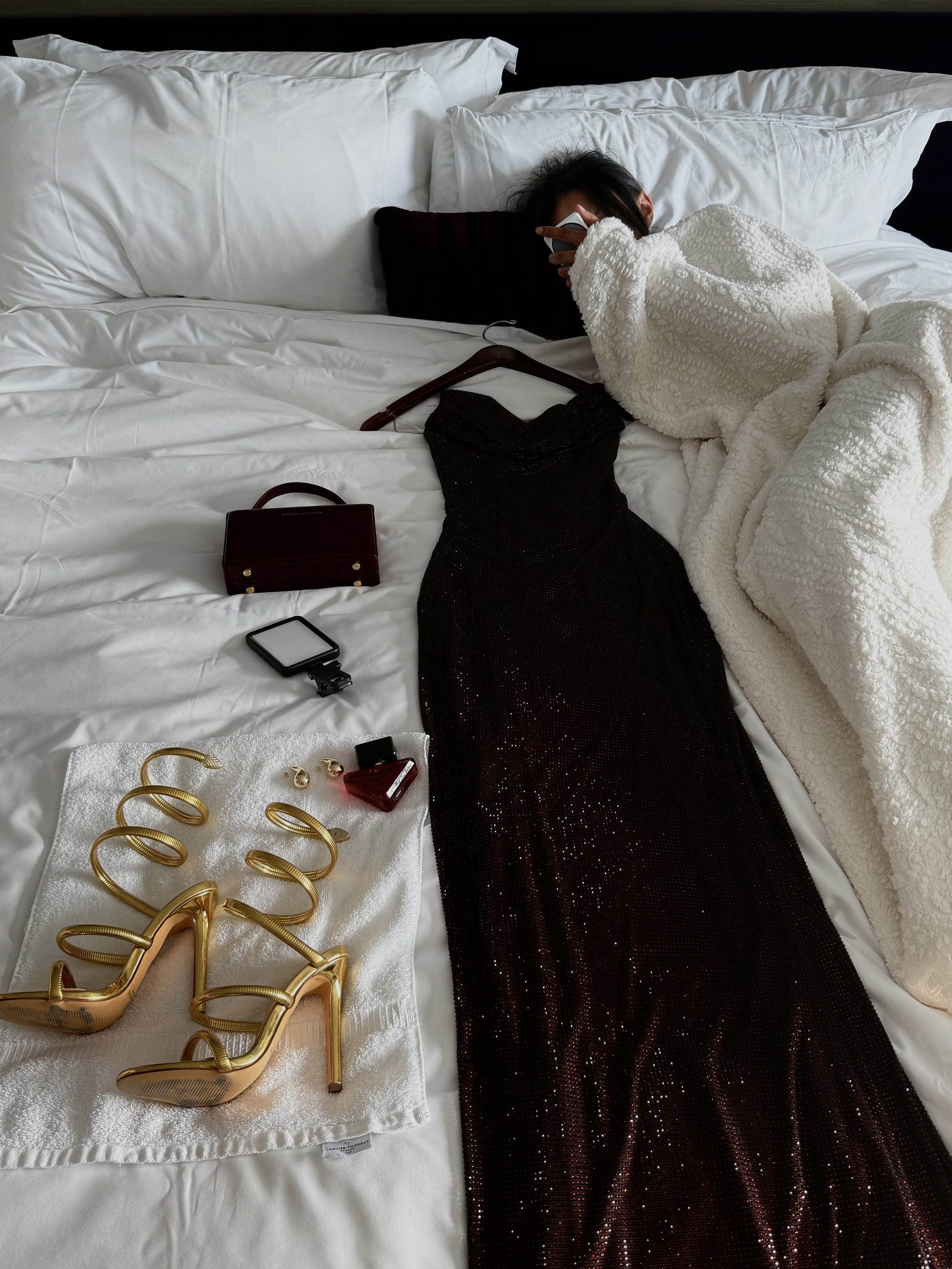 Bree Stephens in bed holding a phone beside a black sequined dress, gold high heels, jewelry, handbag, and white coat.