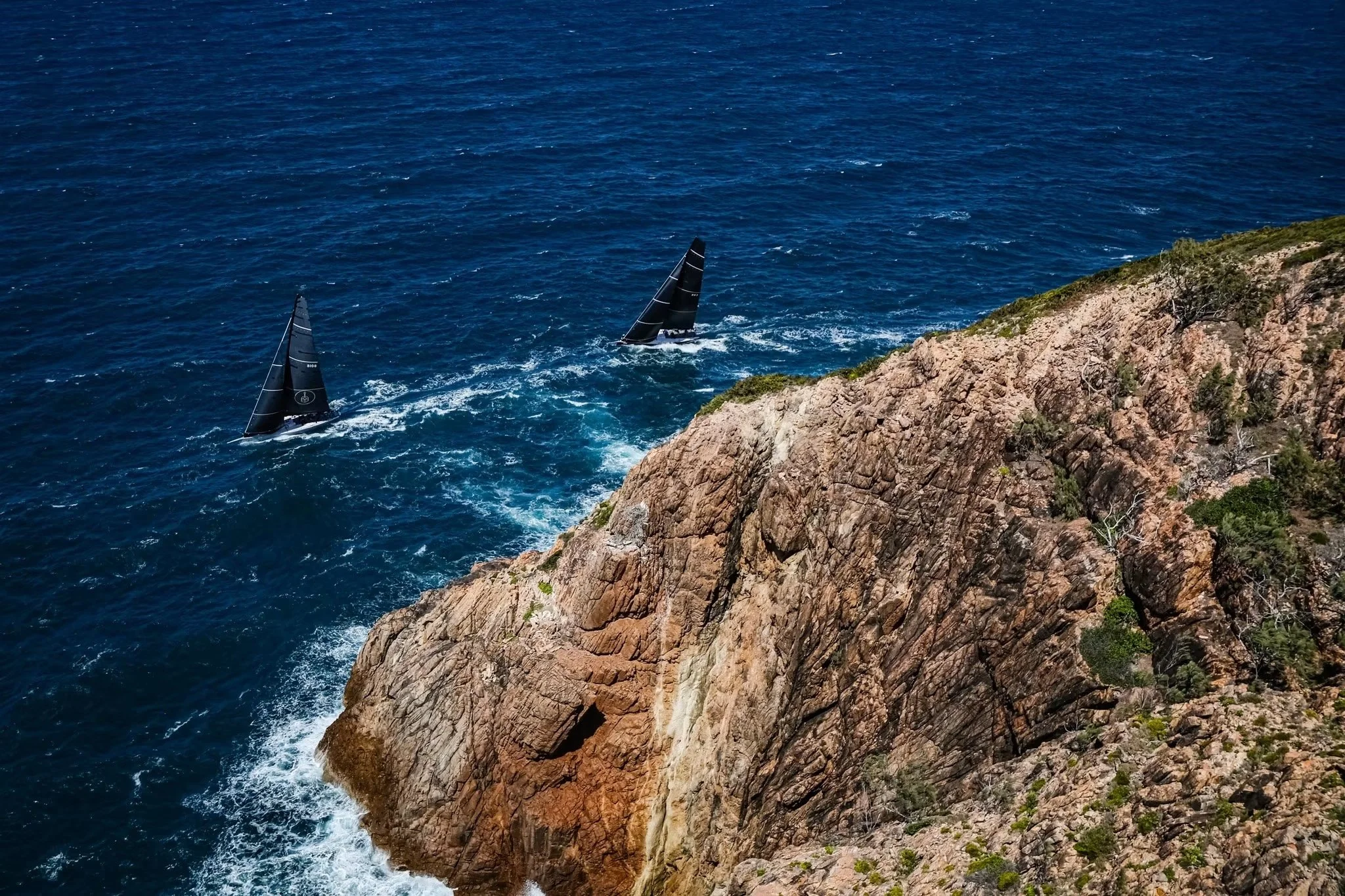 Two sailboats with black sails sailing near rocky coastline on blue ocean under clear sky.