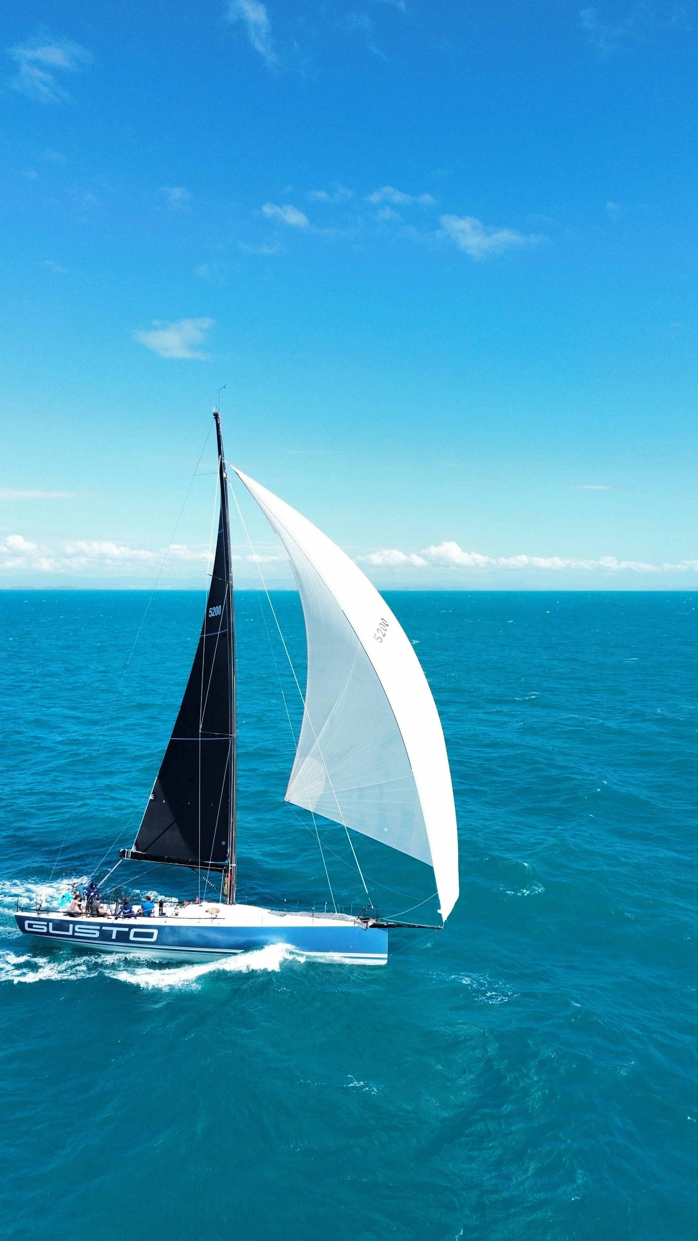 A sailboat named GUSTO sailing on the ocean with bright blue sky and scattered clouds.