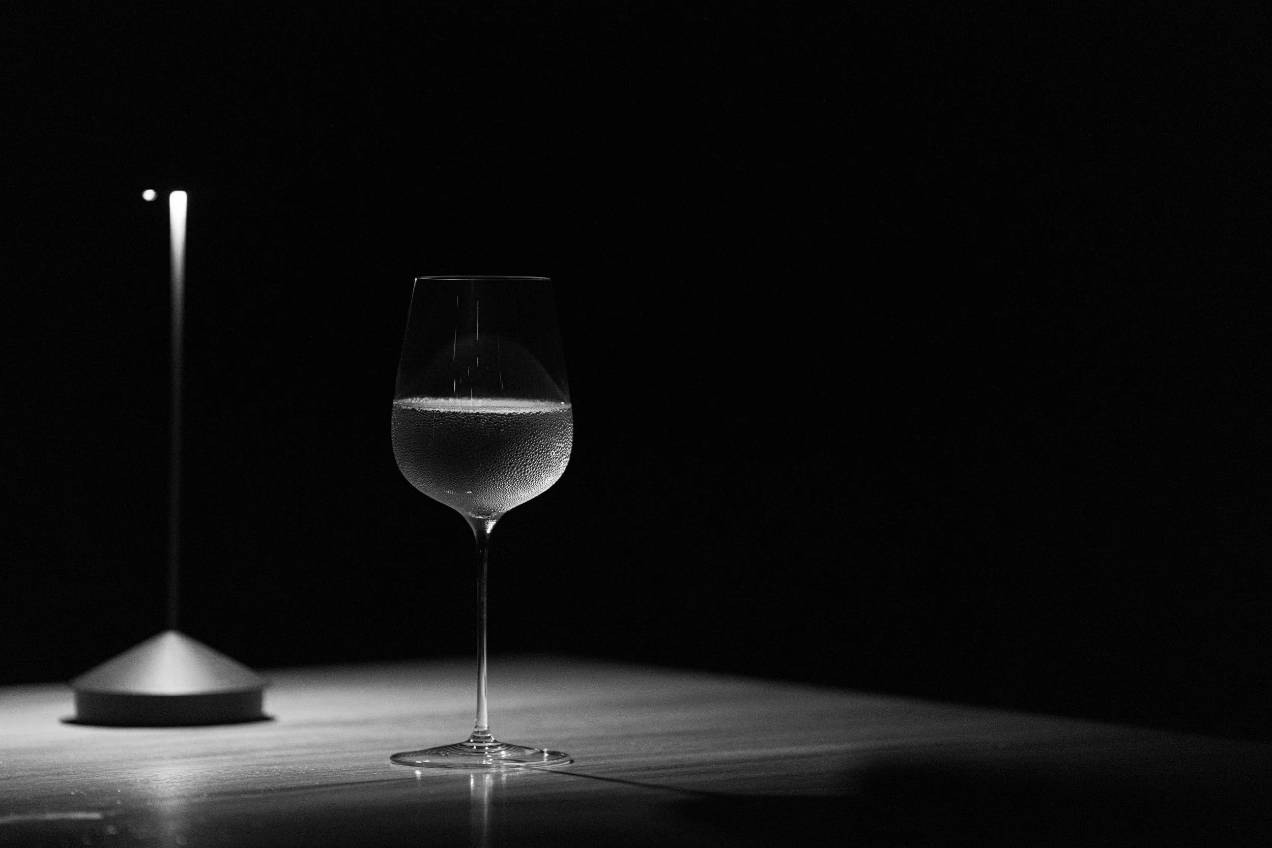 Black-and-white photograph of a wine glass filled with sparkling wine on a reflective table beside a modern lamp.