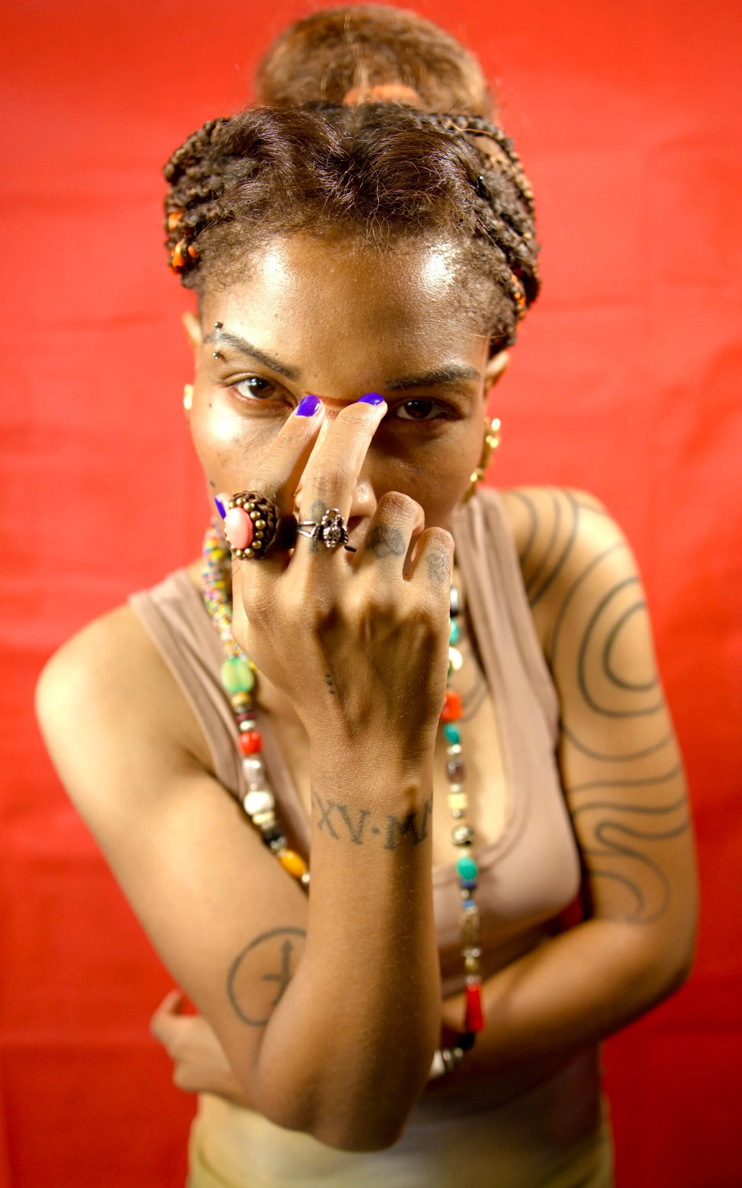 A person with a tattooed arm and multiple rings on their fingers, covering one eye with fingers that have purple nail polish, standing against a red background, wearing layered necklaces and styled hair.
