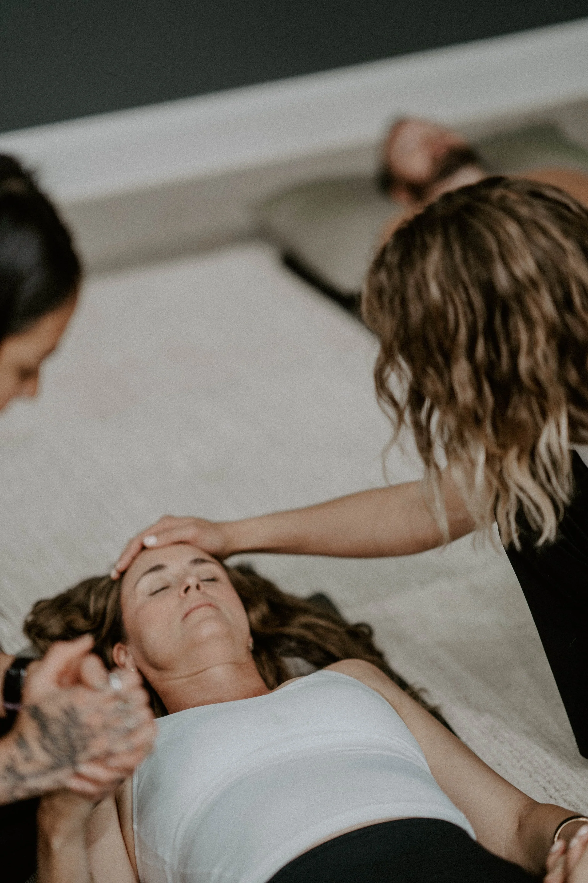 A woman with brown curly hair lying on her back on a white towel, receiving a facial or massage from another woman with long wavy hair. The woman lying down has her eyes closed, appears relaxed, and is wearing a white tank top.