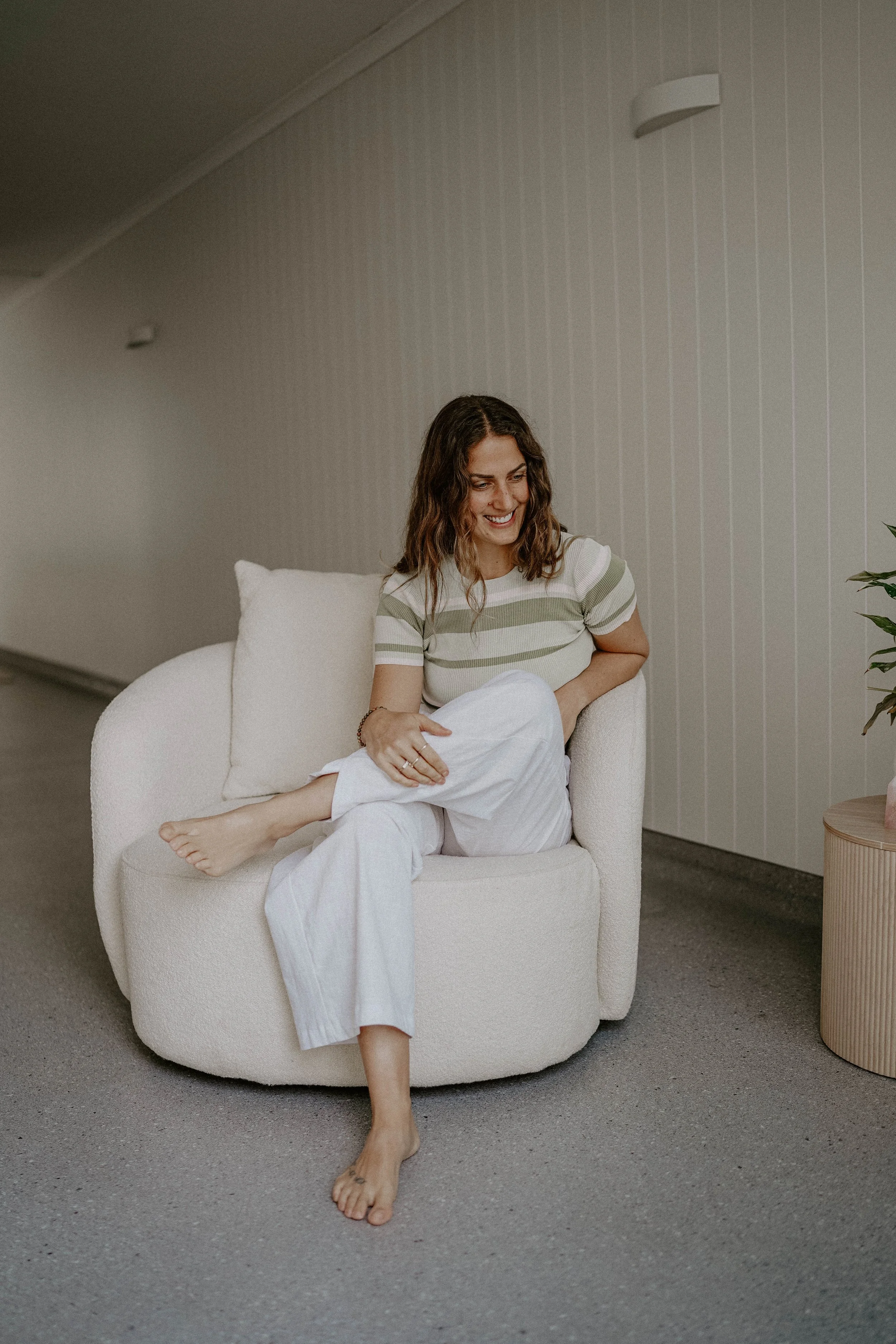 A woman sits on a white upholstered chair in a room with light-colored walls, barefoot, with one leg crossed over the other. She is wearing a short-sleeved, striped shirt and white pants, smiling while looking down.