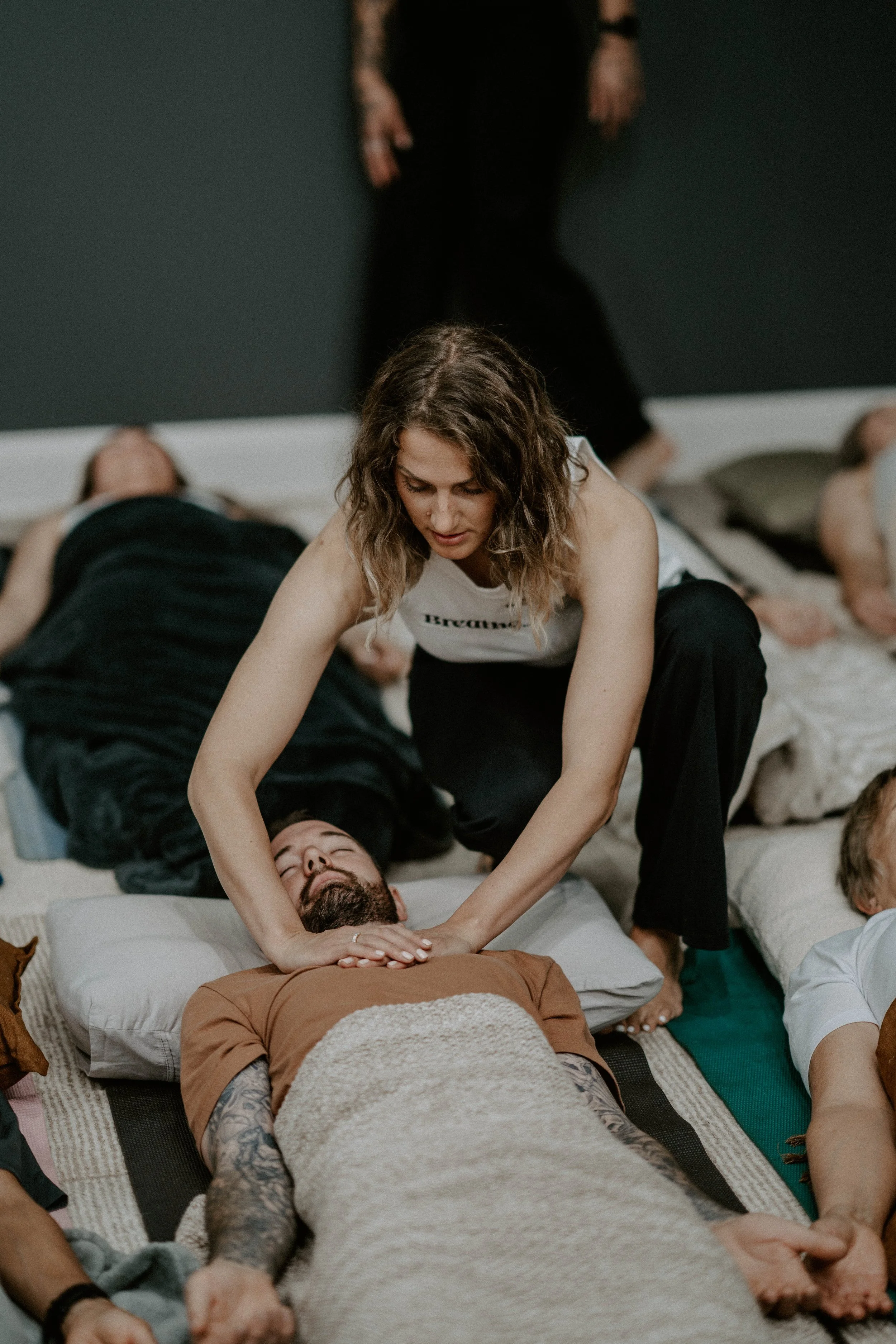 A woman practicing reiki or energy healing on a man lying on a mat, with other people resting on mats in the background.