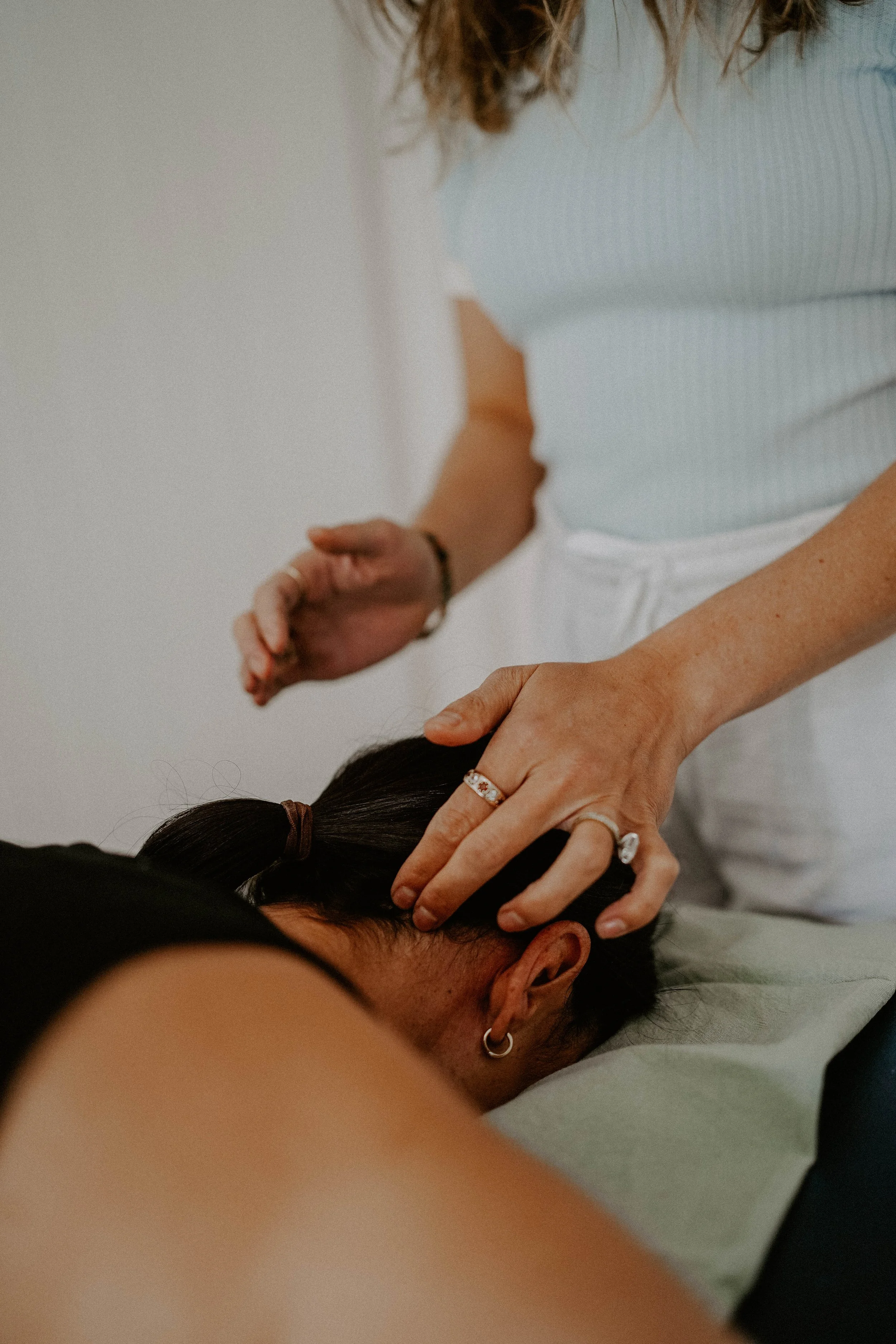 A person receiving a head massage from a practitioner. The practitioner’s hands are on the person's head, and the person is lying down with dark hair tied back. The massage is taking place indoors.
