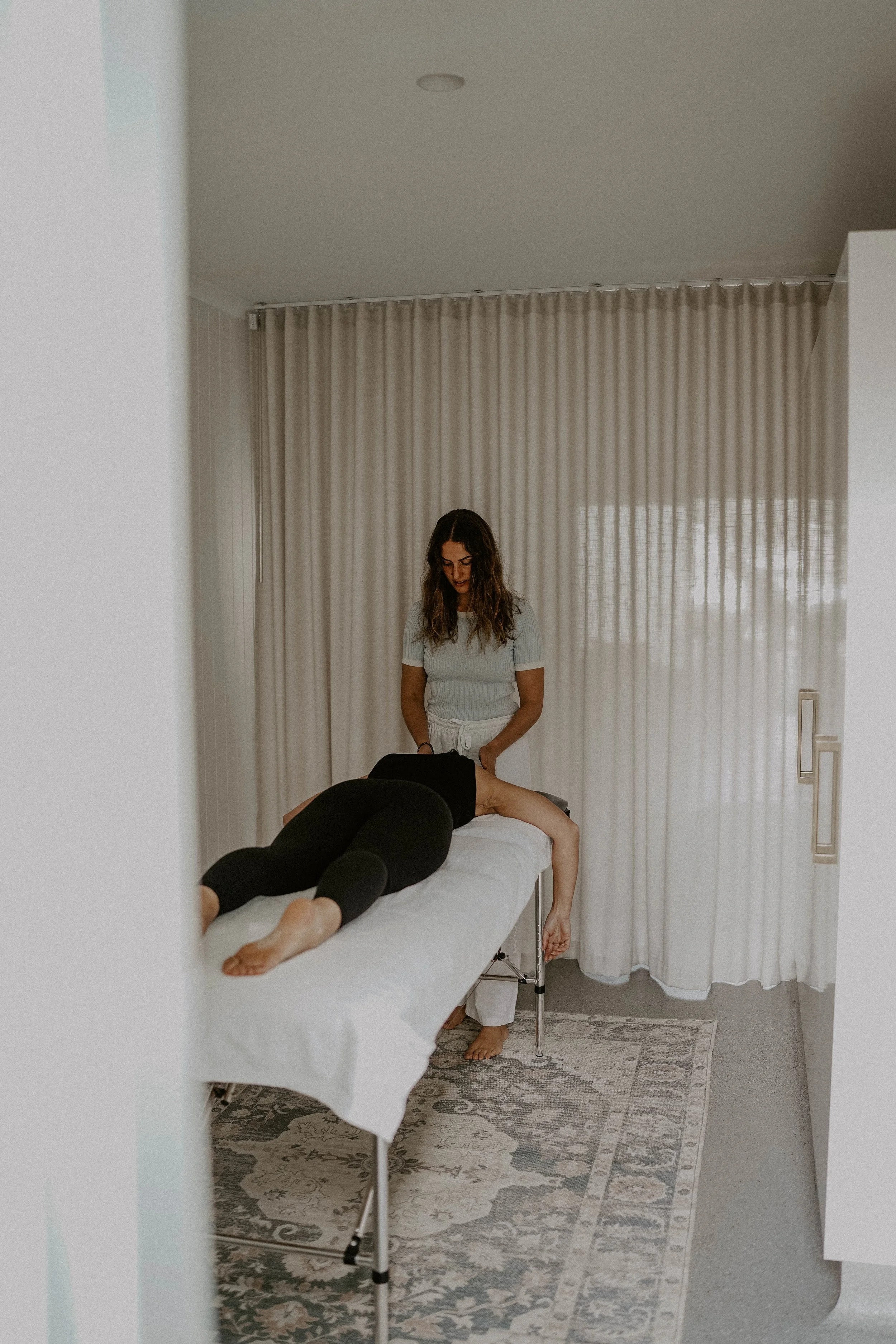A woman receiving a massage from a massage therapist in a room with beige curtains and a patterned rug.