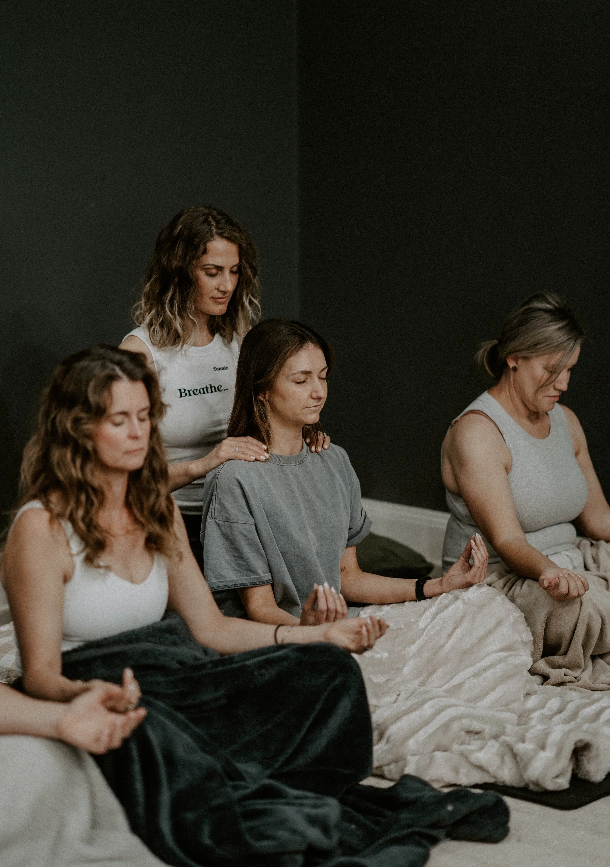 Group of women practicing meditation in a peaceful room, sitting cross-legged with eyes closed and hands resting on knees, some with prayer mudras, surrounded by soft lighting and a cozy blanket on the floor.