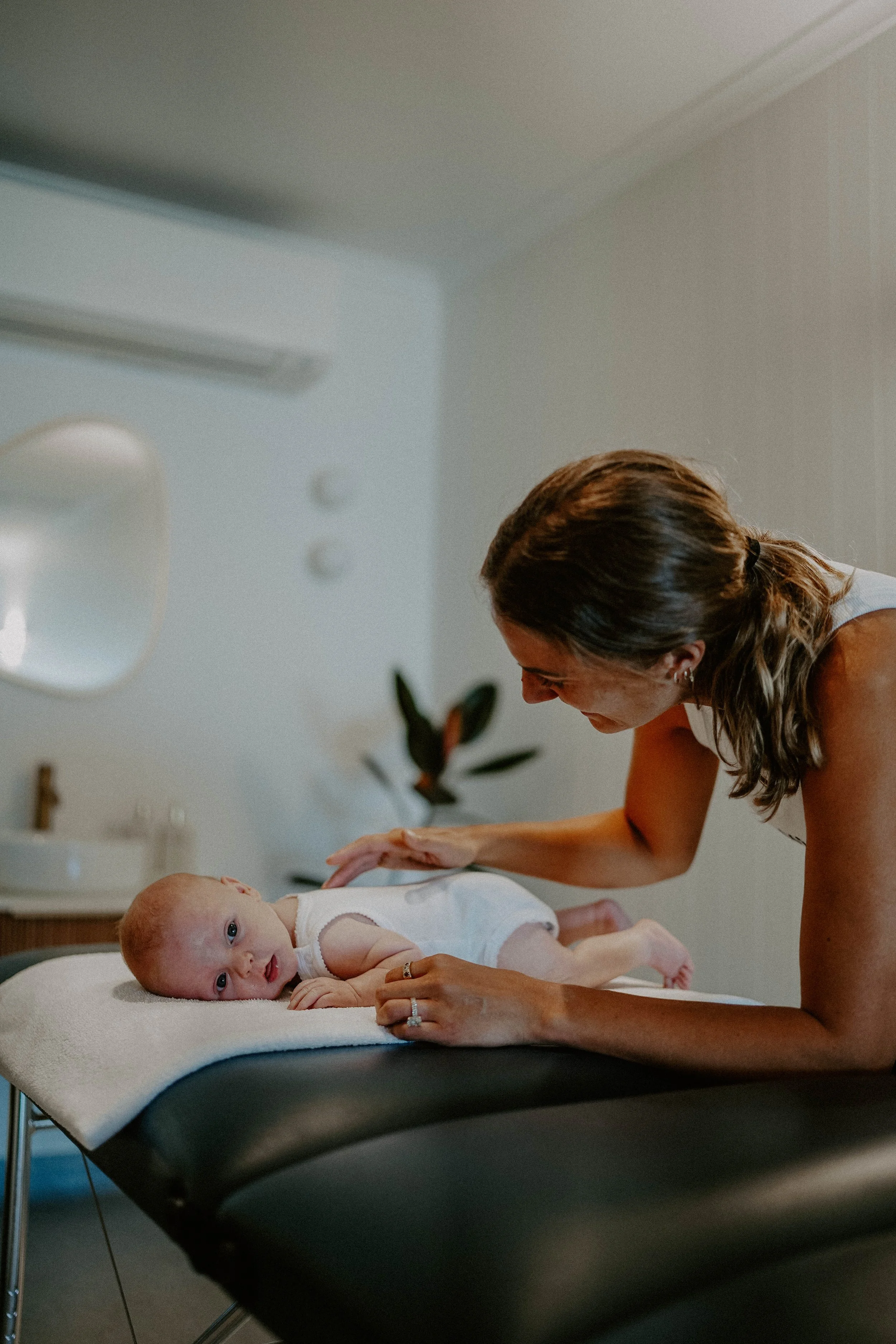 A woman giving a gentle massage to a baby lying on a changing table in a brightly lit room.