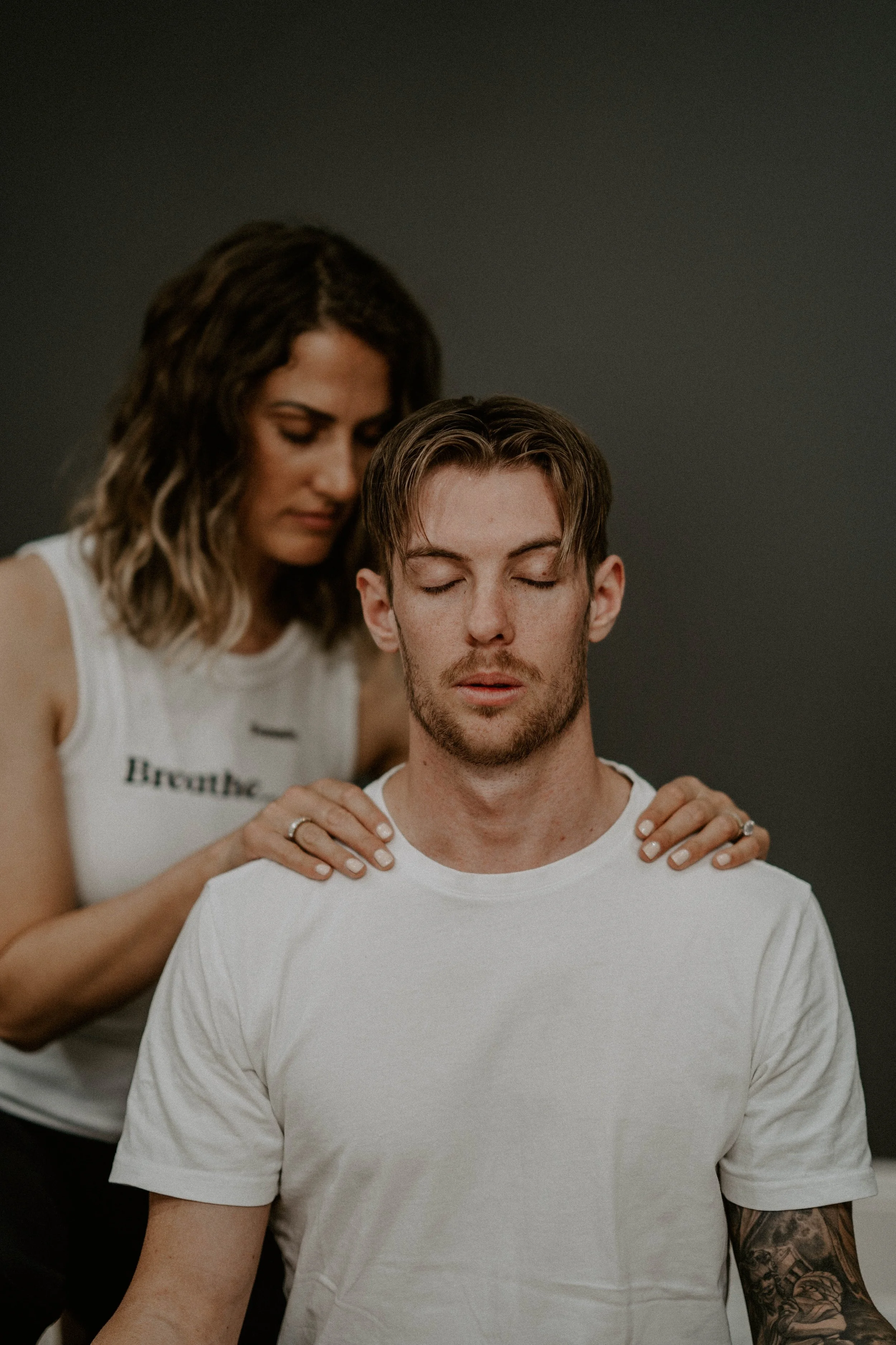 A woman with wavy brown hair leaning over a seated man with closed eyes, resting her hands on his shoulders in a calming gesture.