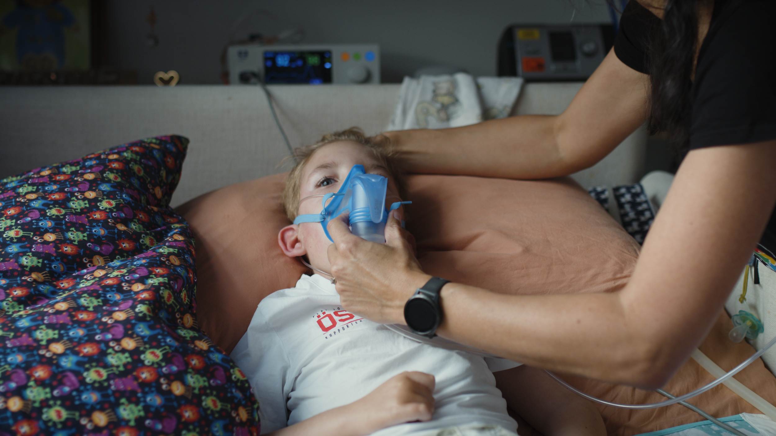 A parent gently holds a blue breathing mask over a young boy’s face as he lies on a patterned pillow, monitors on the shelf behind.