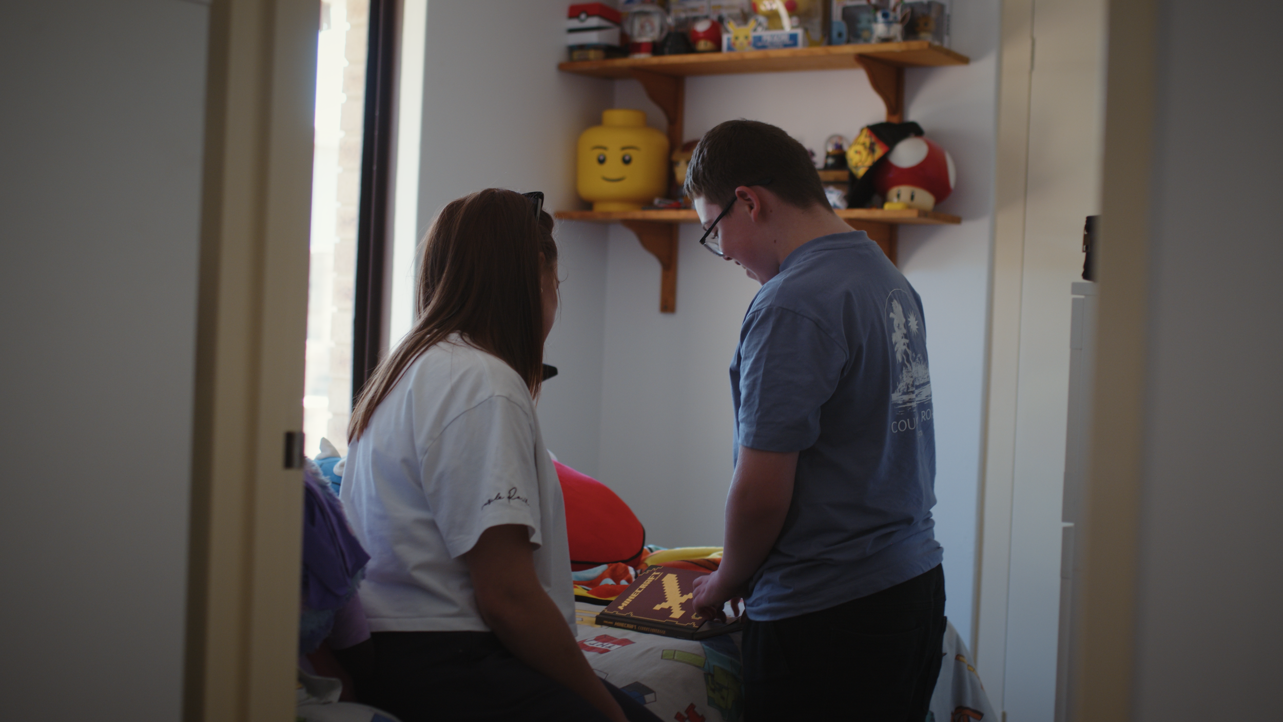 A parent and her teenage son in his bedroom, seen from behind, with toys and a Lego head on the shelves above.