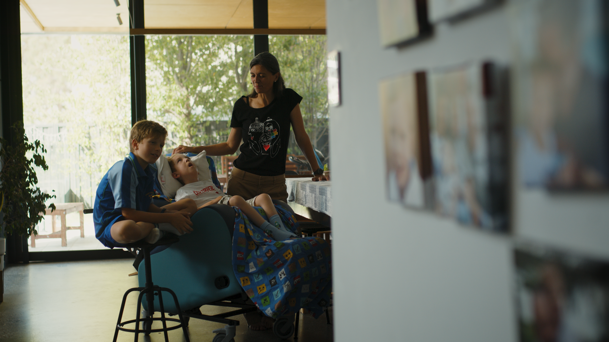 A woman leans over a child in a supported chair while a boy sits close by, in a sunny room with family photos on the wall.