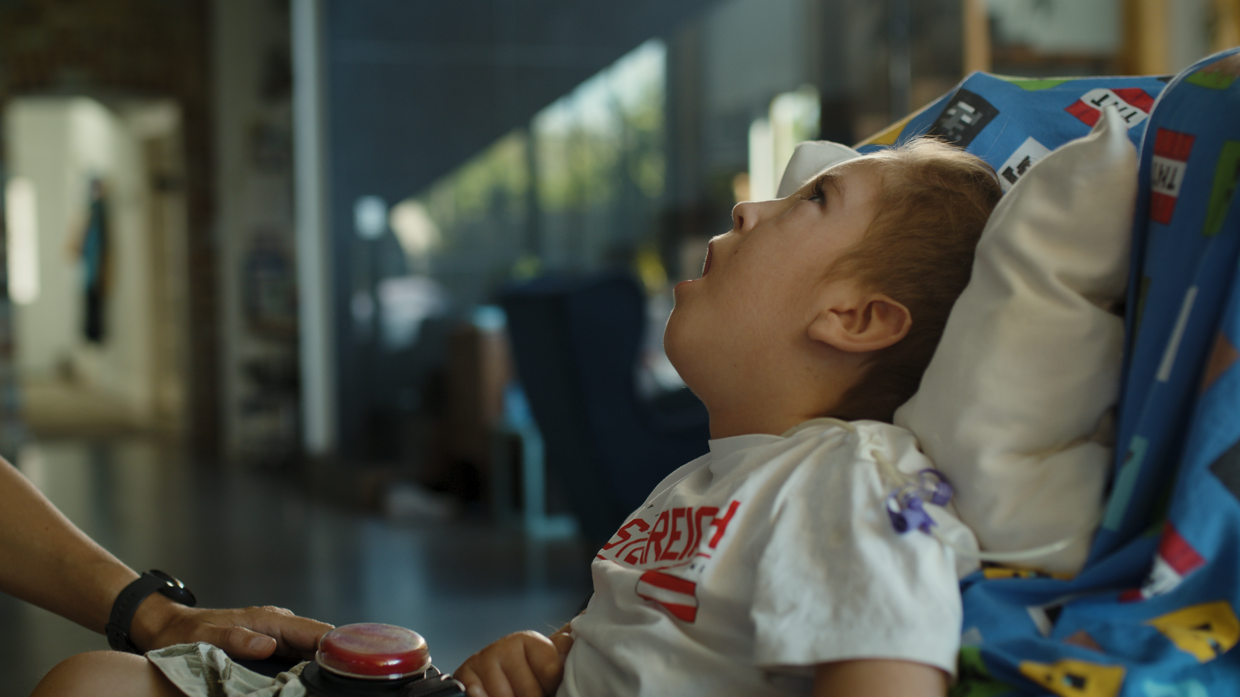 A boy leans back in a supported seat by bright windows, looking up and away.