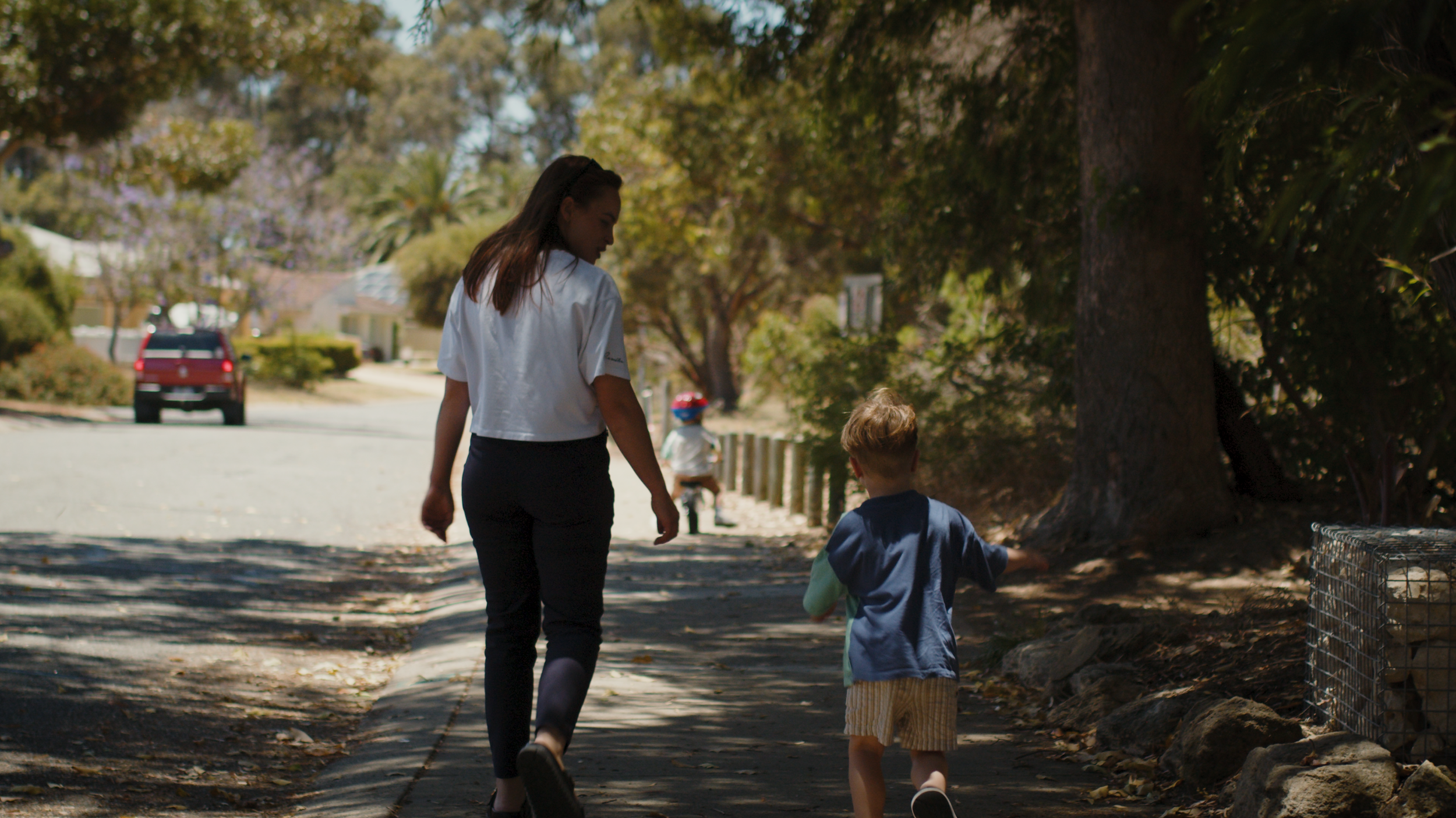 A mother walks along a leafy suburban footpath with two young children.