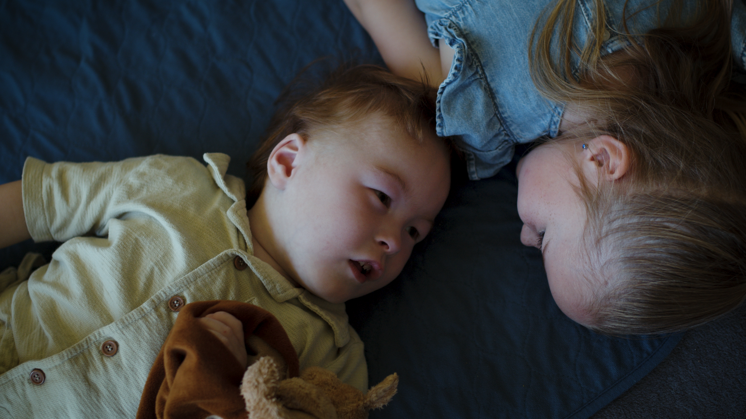 A toddler and a young girl lie side by side on a blue blanket, faces close together.