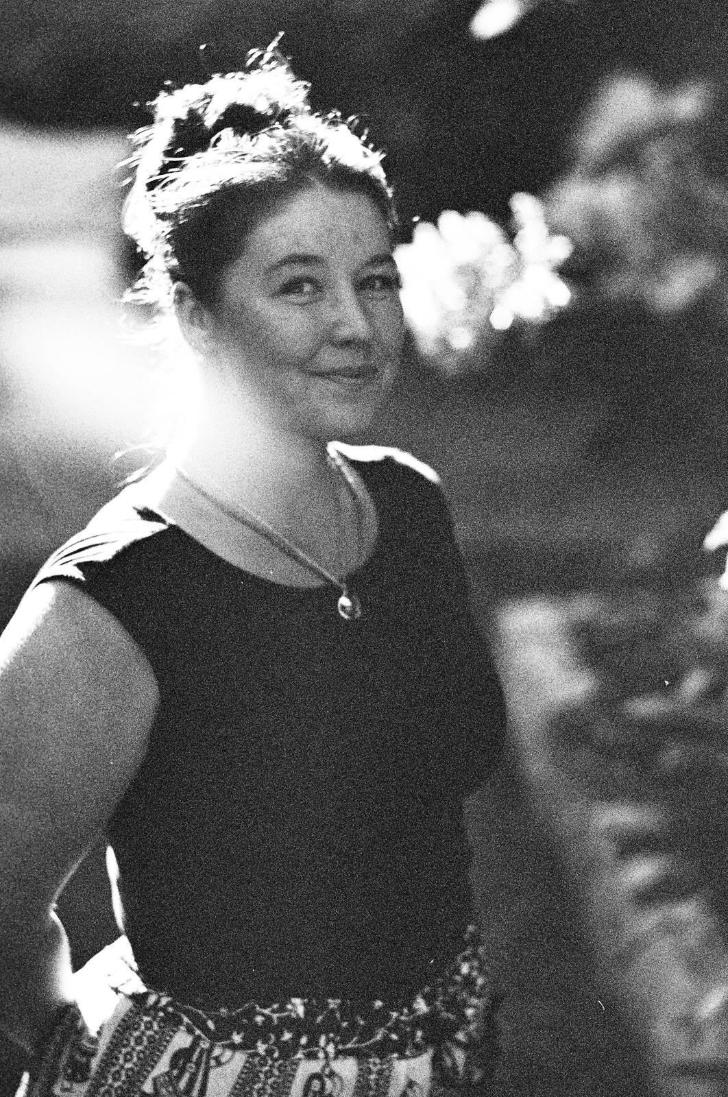 Black and white photo of a smiling woman with short hair, wearing a dark top, a necklace, and patterned pants, outdoors at night with blurred lights in the background.
