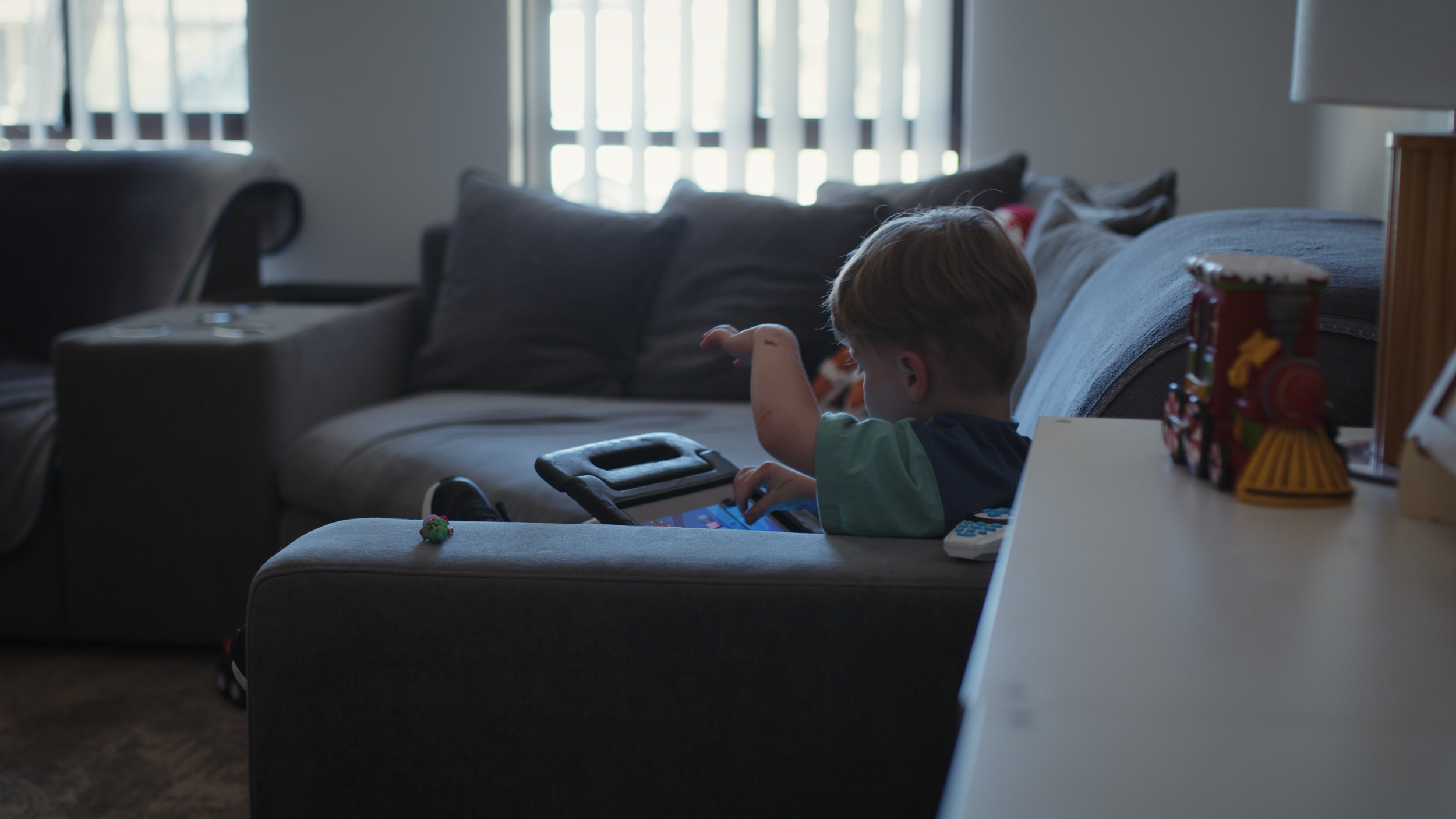 A young boy kneels on the couch in a softly lit living room, looking out the window.