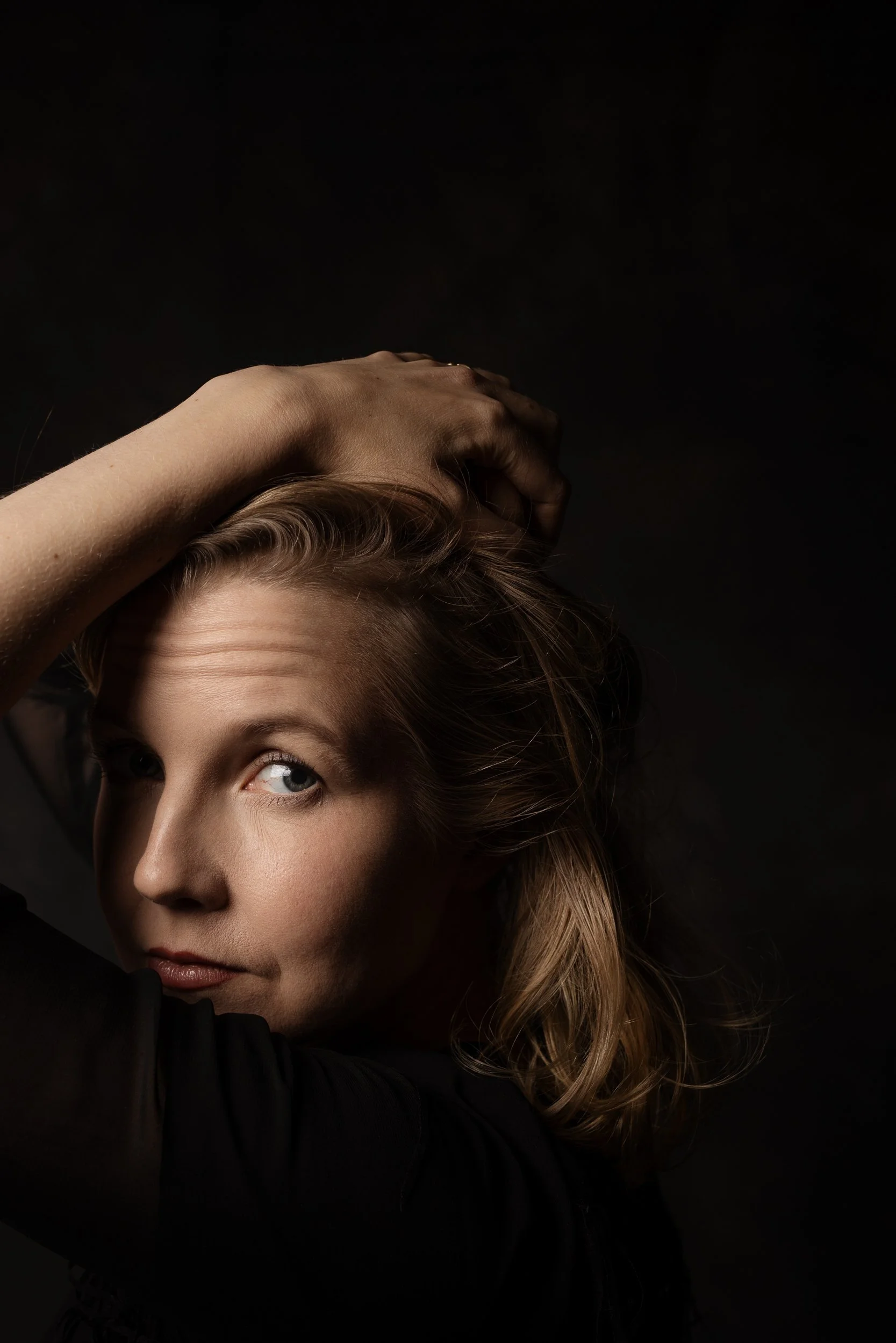 Hämärä muotokuva naisesta, joka katsoo sivulle käsi pään päällä mustaa taustaa vasten.