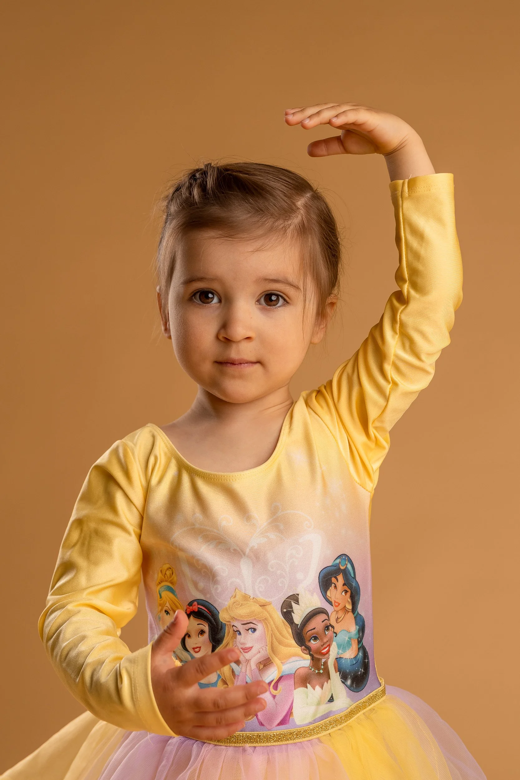 Pieni tyttö keltaisessa prinsessakuvioisessa mekossa nostaa kätensä ruskeaa taustaa vasten.