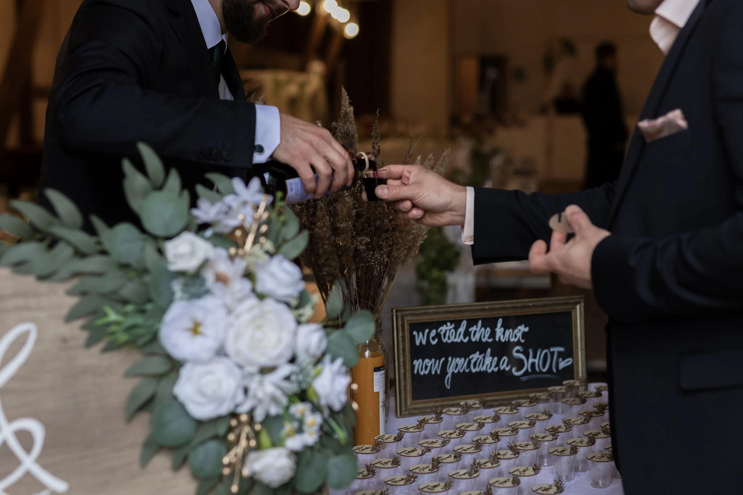 Häissä mies kaataa juomaa vieraalle, taustalla kukka-asetelma ja kyltti “We tied the knot, now you take a shot”.