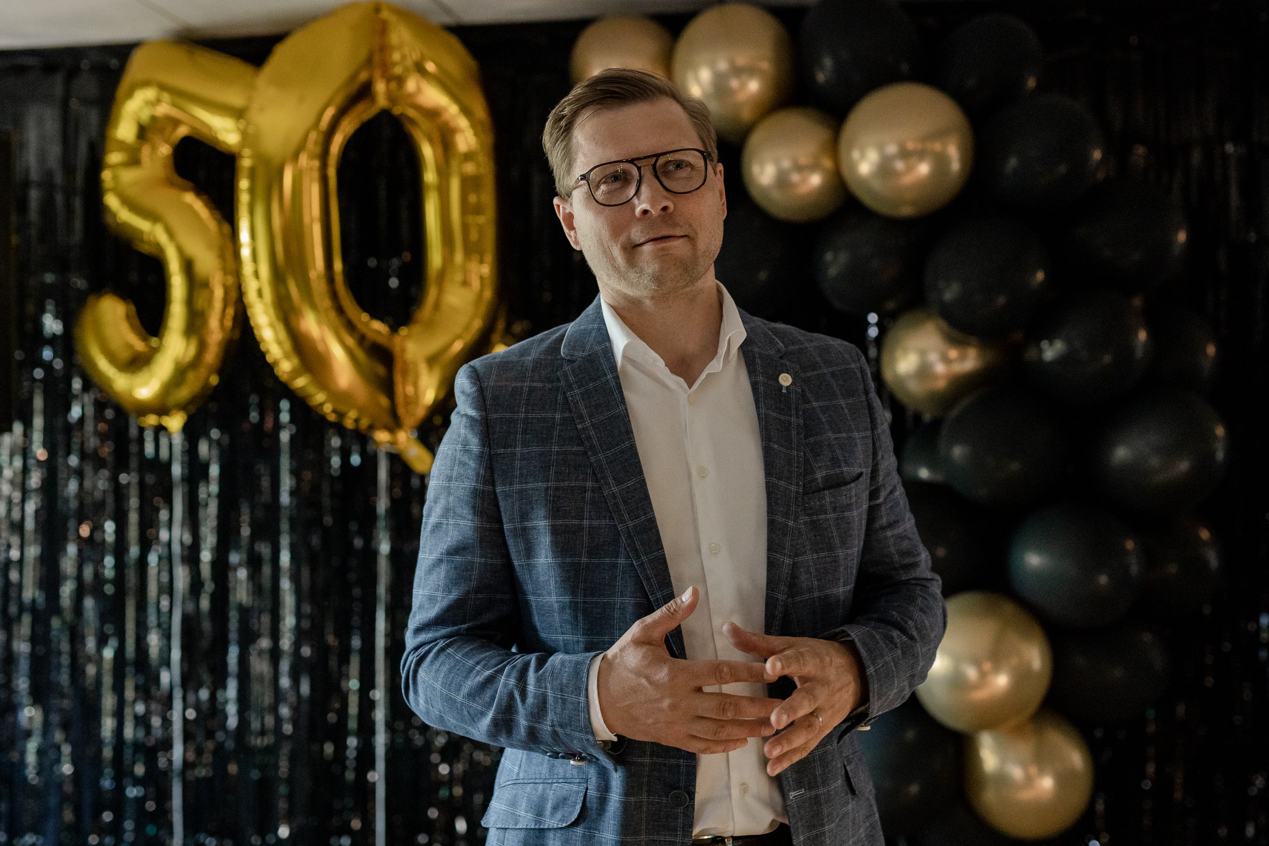 Silmälasipäinen mies ruudullisessa puvuntakissa ja valkoisessa paidassa juhlavasti koristellussa tilassa ilmapallojen edessä.