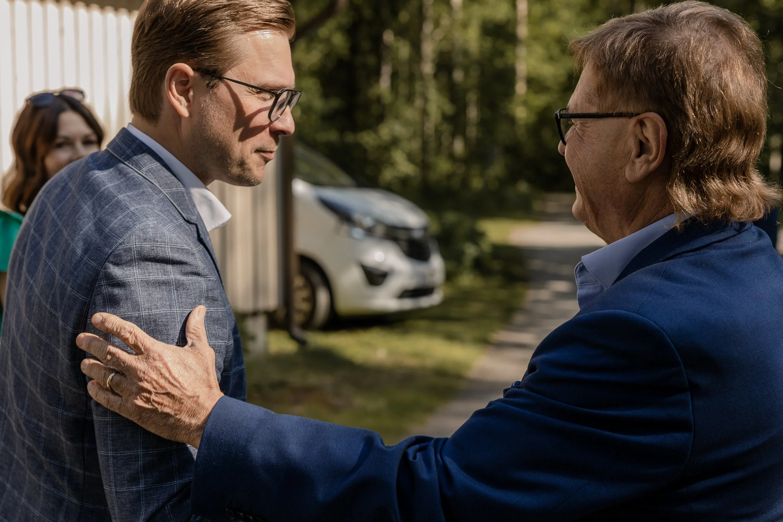 Kaksi silmälasipäistä miestä keskustelee ulkona; iäkäs mies pitää kättään toisen olkapäällä.