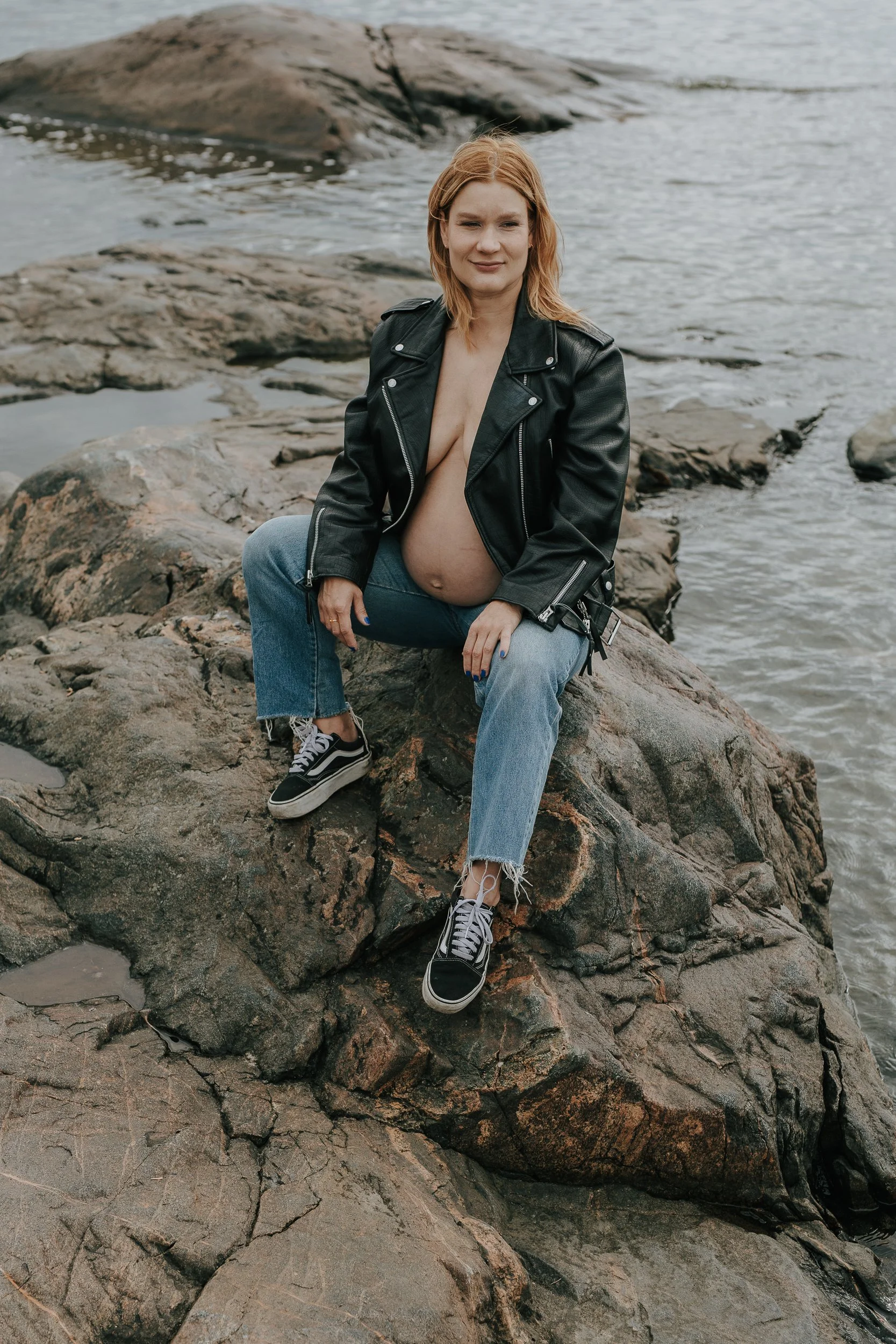 Raskaana oleva nainen istuu kallioilla veden äärellä mustassa nahkatakissa ja farkuissa.